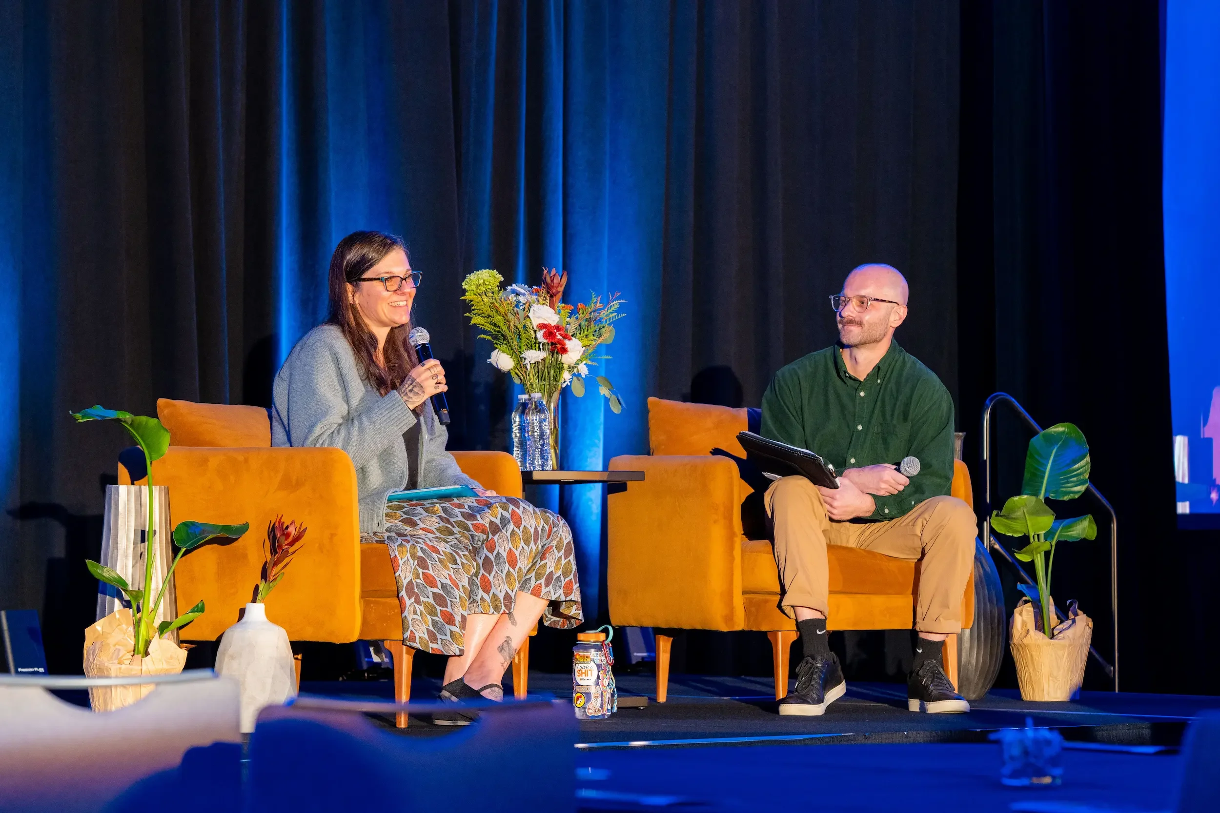 Two people sitting on orange chairs on a stage with dark blue curtains. The woman on the left is speaking into a microphone and smiling, while the man on the right is listening, holding a tablet. There are plants and flowers on the stage.