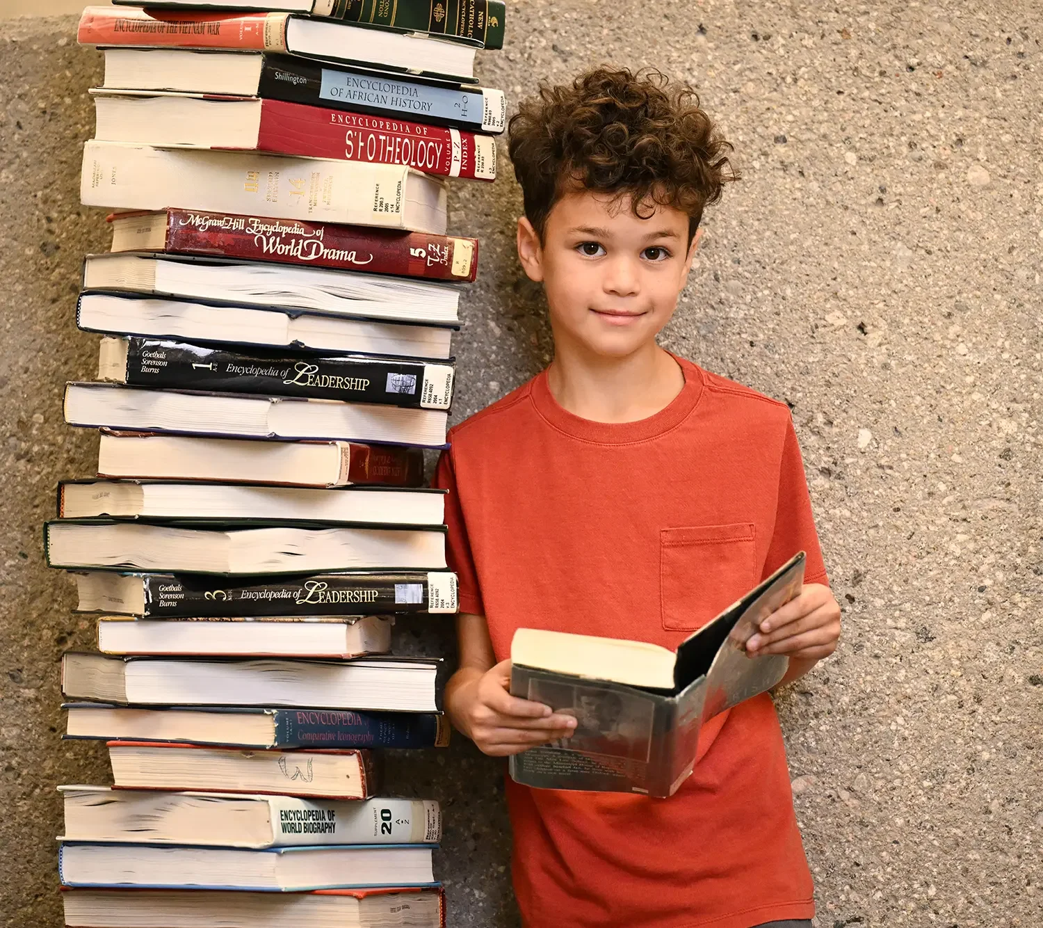A young boy with curly hair and a red t-shirt standing outdoors next to a large stack of books, holding an open book in his hands and smiling at the camera.
