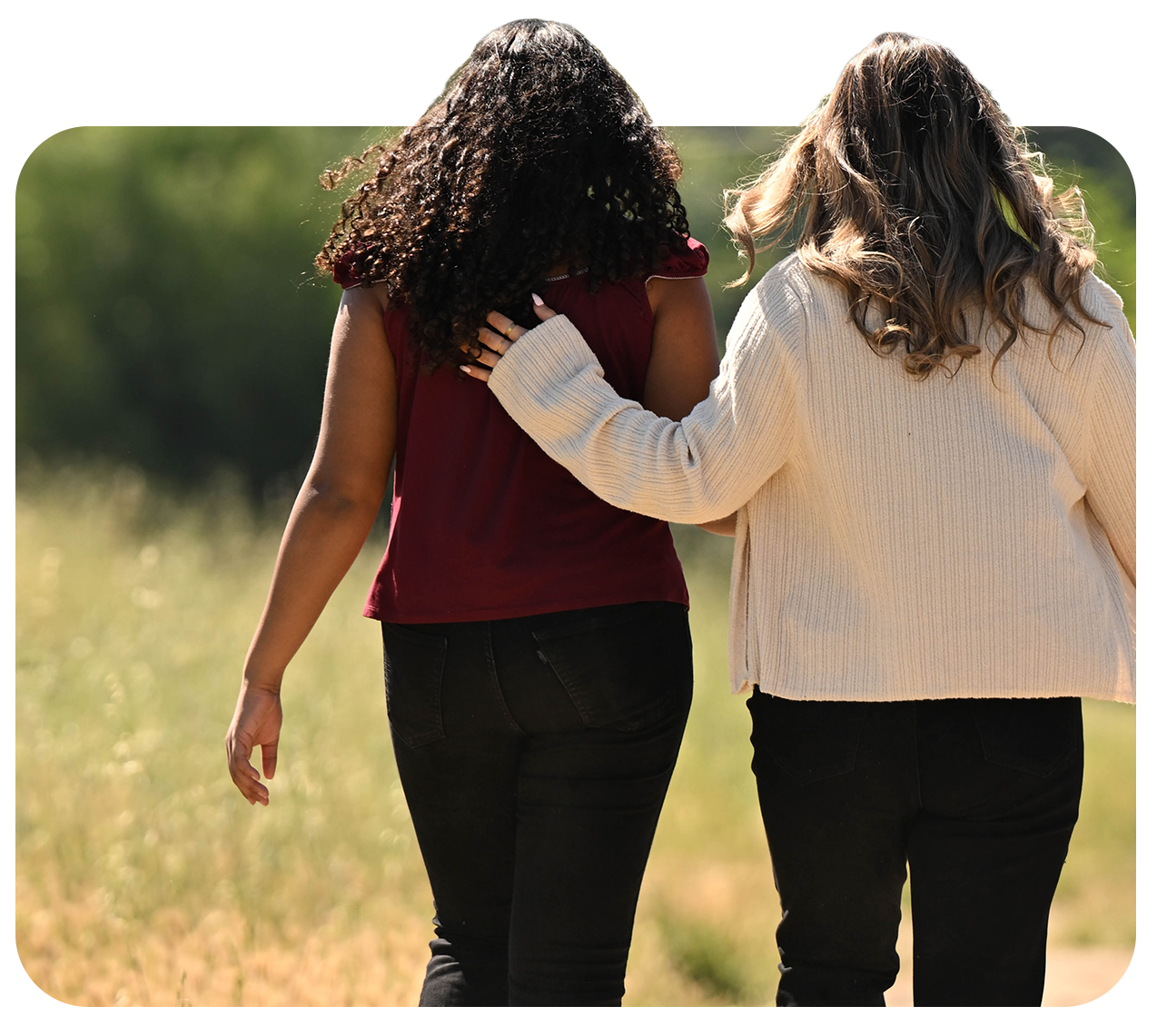 Two women walking together outdoors, one with curly hair wearing a red top, and the other with wavy hair wearing a cream sweater, with the woman in the cream sweater gently touching the other woman's shoulder.
