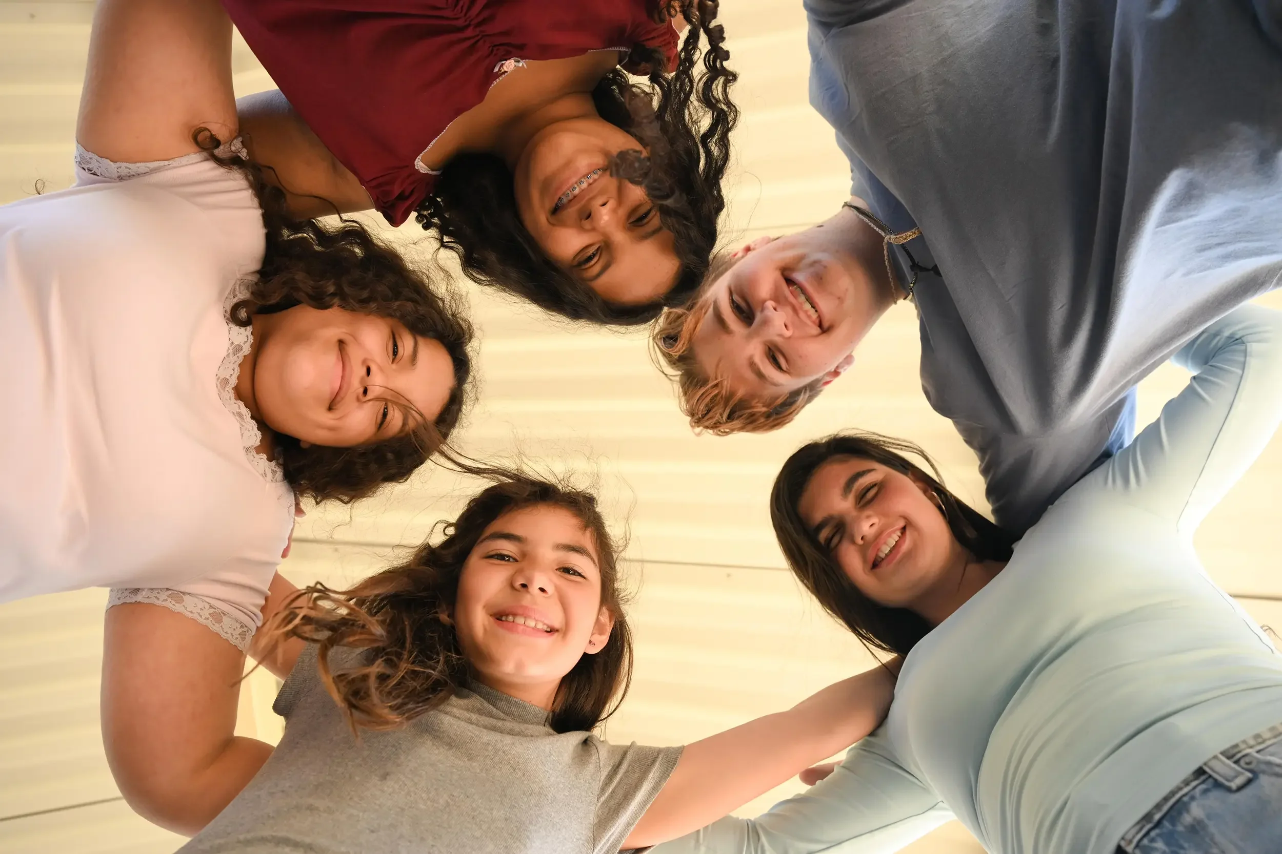 A group of smiling women and girls gathered closely together, looking down at the camera, creating a circle with their heads and arms.