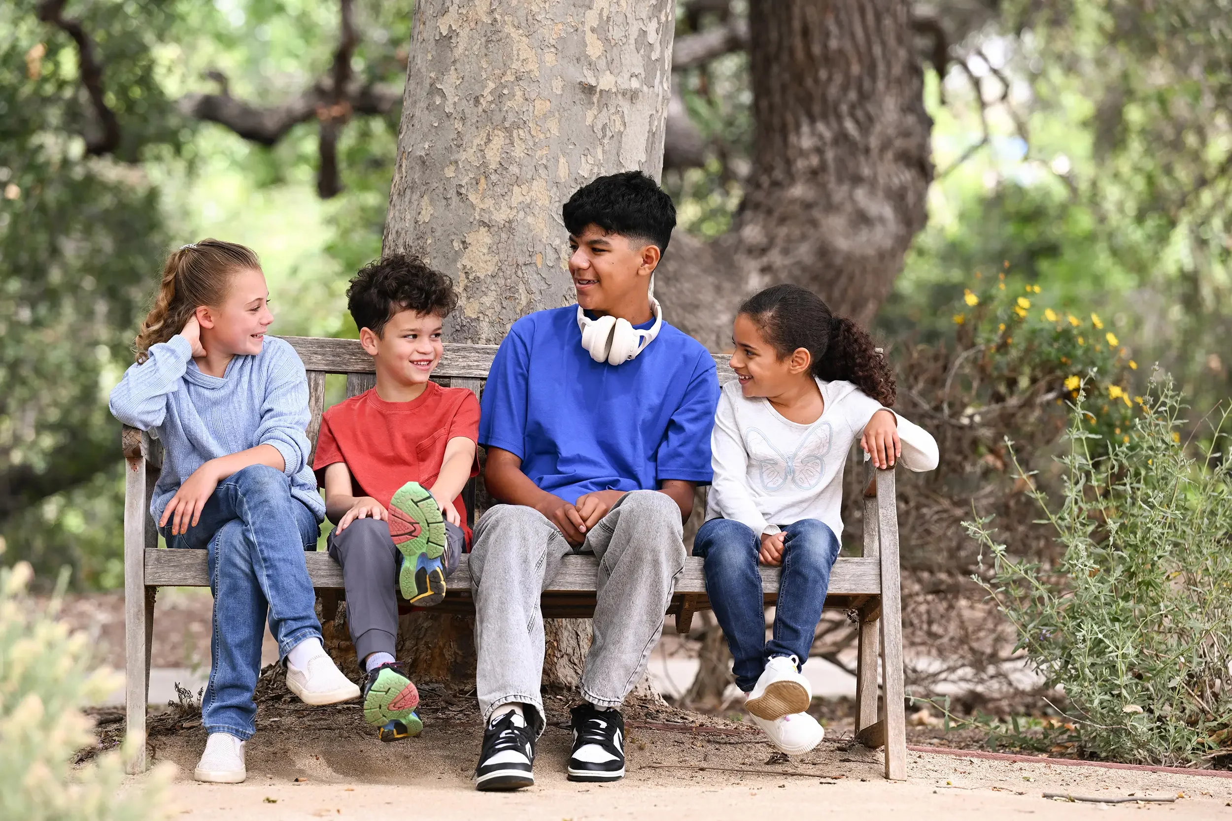 A group of five kids sitting on a park bench in front of a large tree, smiling and talking to each other.