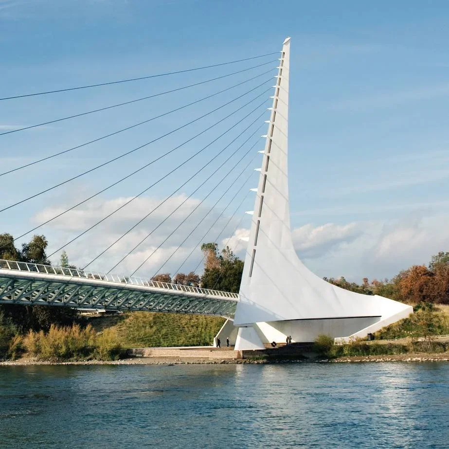 A modern white sail-shaped bridge with cables extending from a tall spire, crossing over a river with trees on the shoreline.