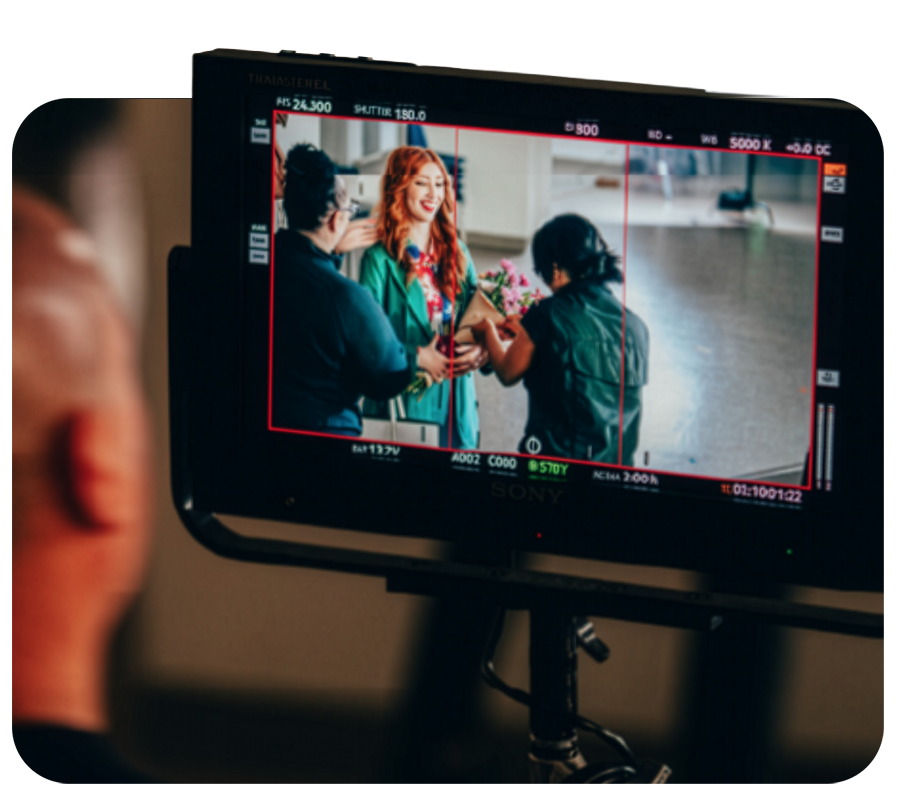 Behind the scenes of a photo shoot, a monitor displays a woman with red hair smiling in a green jacket, receiving flowers from two women. A person in the foreground observes the screen.
