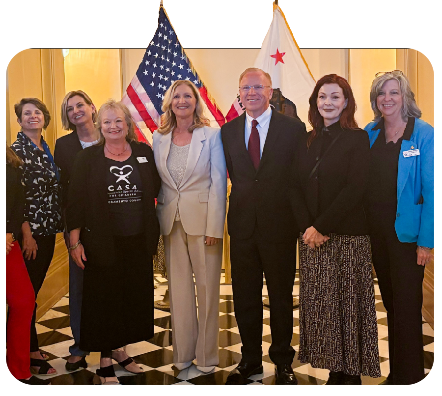 Group of seven women and one man standing together in front of a US flag and a California flag, indoors, with a checkered black and white floor.