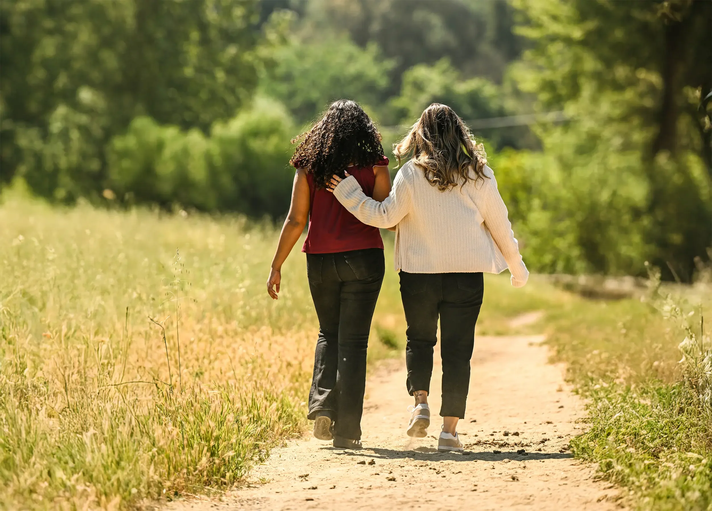 Two women walking on a dirt path through a green, sunny forest, with one arm around each other's shoulders.