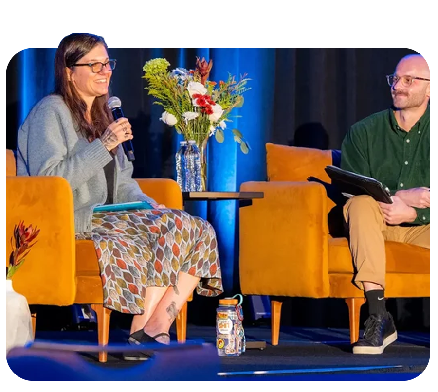A woman and a man sitting on yellow armchairs on a stage, engaged in a conversation. The woman is holding a microphone and smiling, while the man is holding a tablet. There is a table with a flower vase between them.