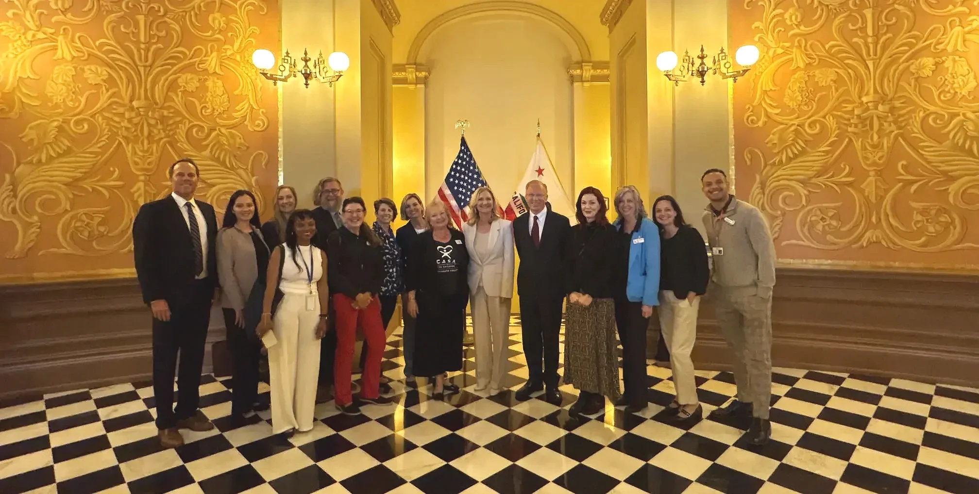 A group of people standing together indoors, in front of a wall with ornate gold decoration and two flags (American and California) behind them. The group includes men and women dressed in business or casual attire, smiling for the photo.