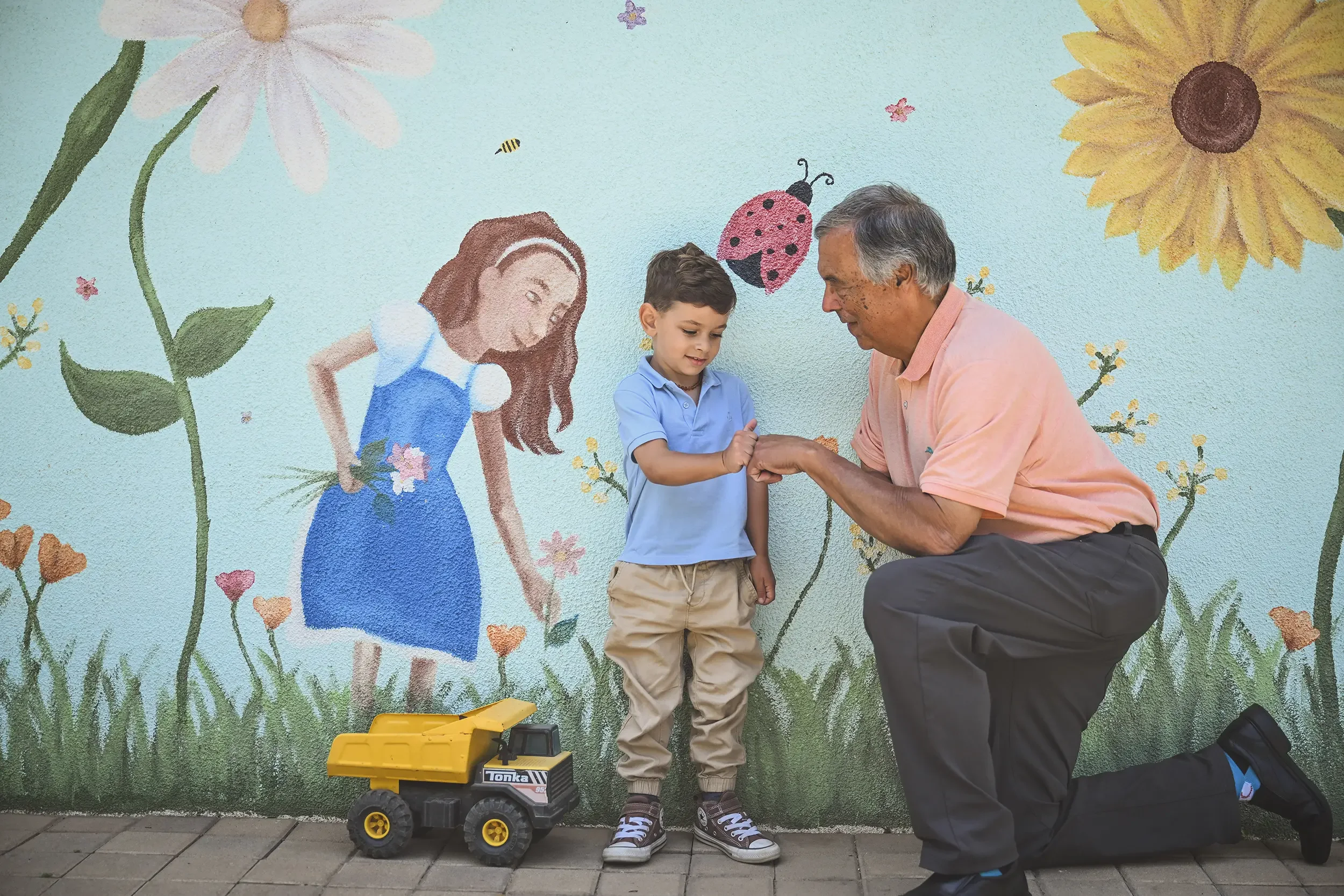 A man kneeling and a young boy standing in front of a painted wall with flowers and insects, exchanging a handshake. There is a yellow toy truck on the ground next to them.