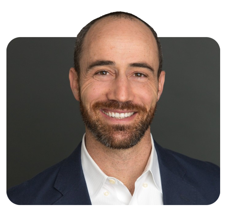Headshot of a smiling man with a beard and short hair, wearing a navy blazer and white shirt against a dark background.