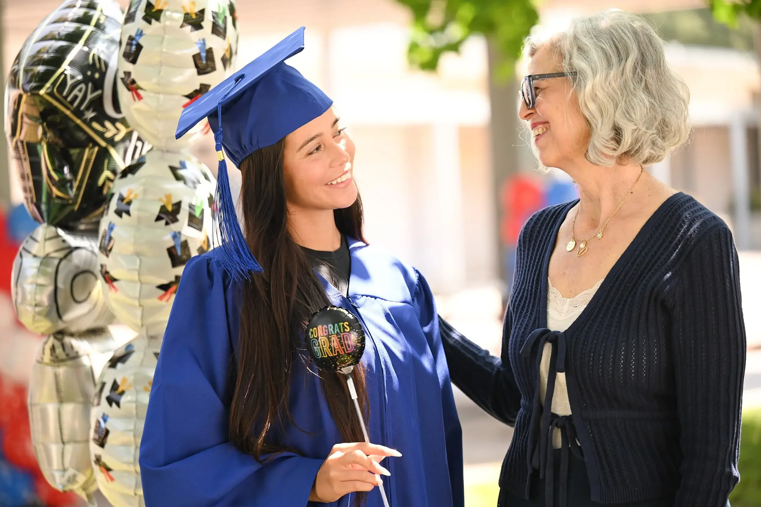 A young woman in a blue graduation cap and gown smiling at an older woman, possibly her mother, outside on a sunny day, with gold and black balloons in the background.