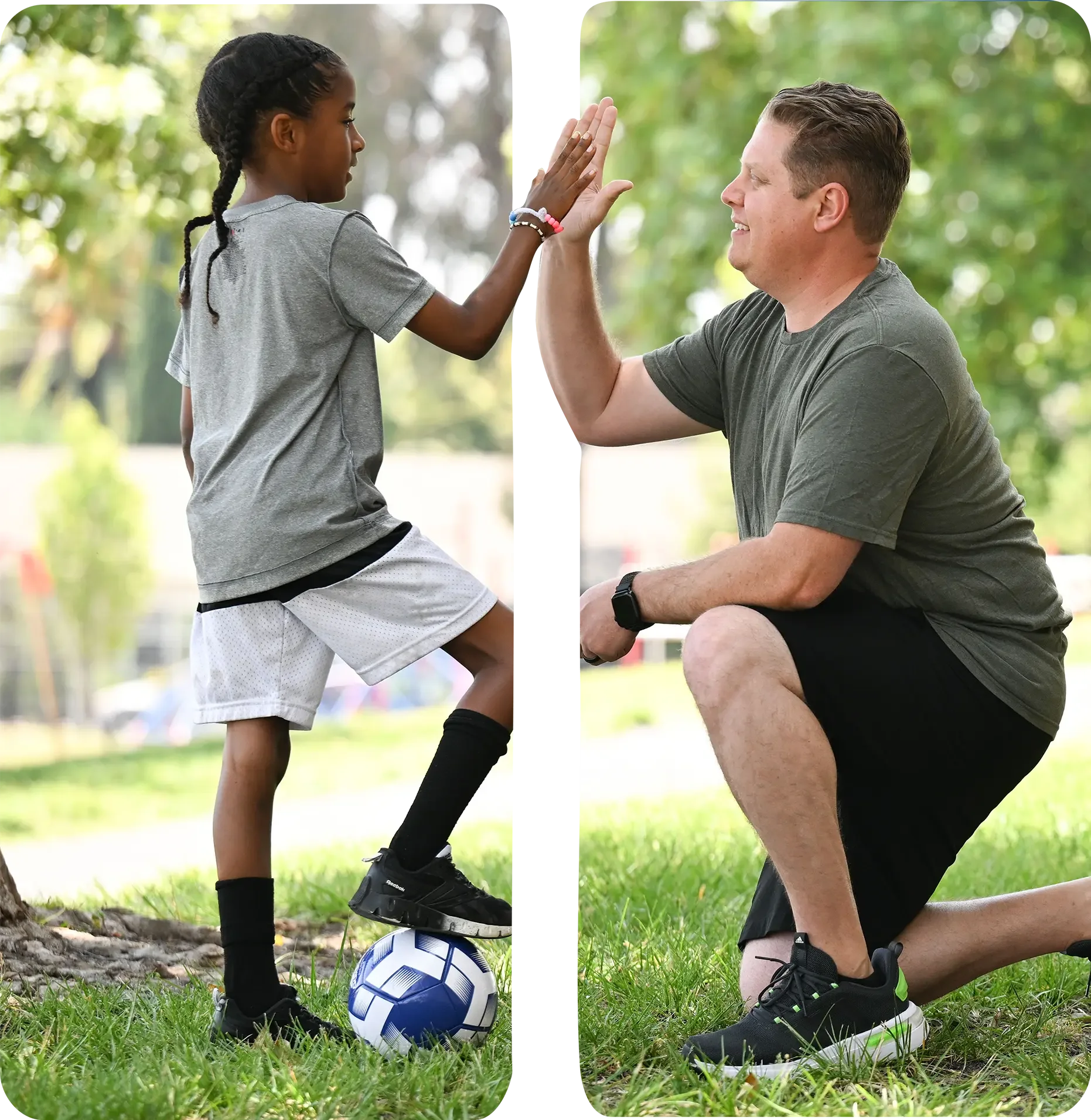 A young girl in gray sportswear and black socks high-fives a man in gray athletic clothing while kneeling on the grass, with a soccer ball at their feet in a park with trees in the background.