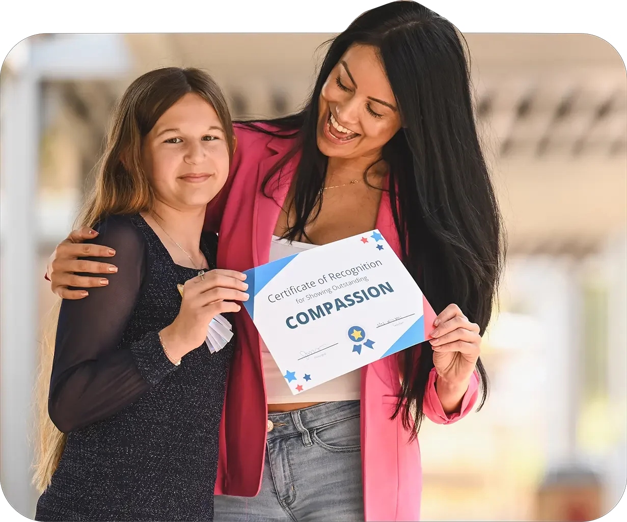 A woman with long dark hair and a pink blazer shows a certificate of recognition for showing compassion to a smiling young girl with long blonde hair, wearing a black top, in an indoor setting.