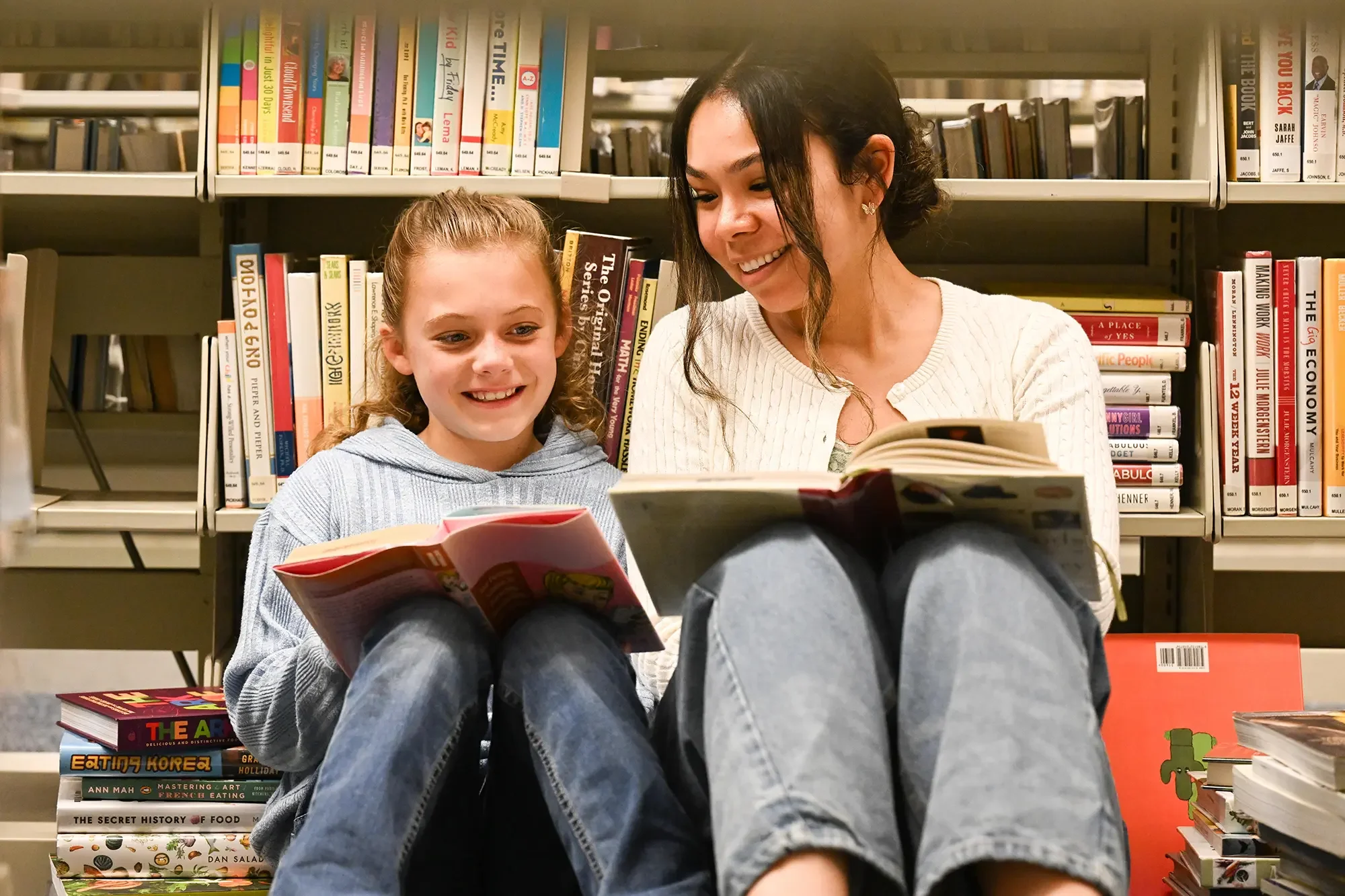 A woman and a young girl sitting on the floor in a library surrounded by shelves of colorful books, smiling and reading books together.