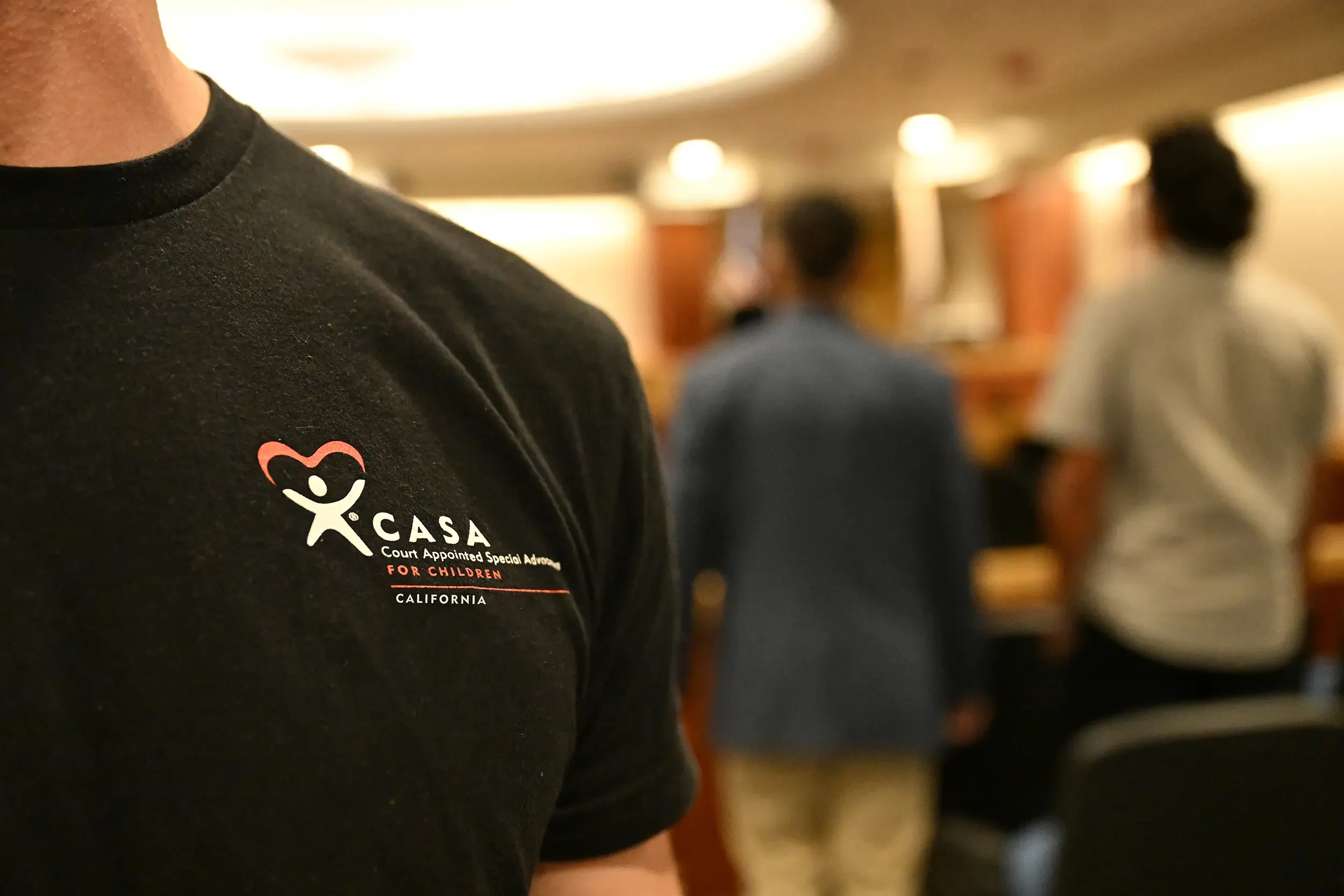 Close-up of a person wearing a black shirt with a logo for CASA, an organization for court-appointed special advocates for children, with a blurred background of other people.