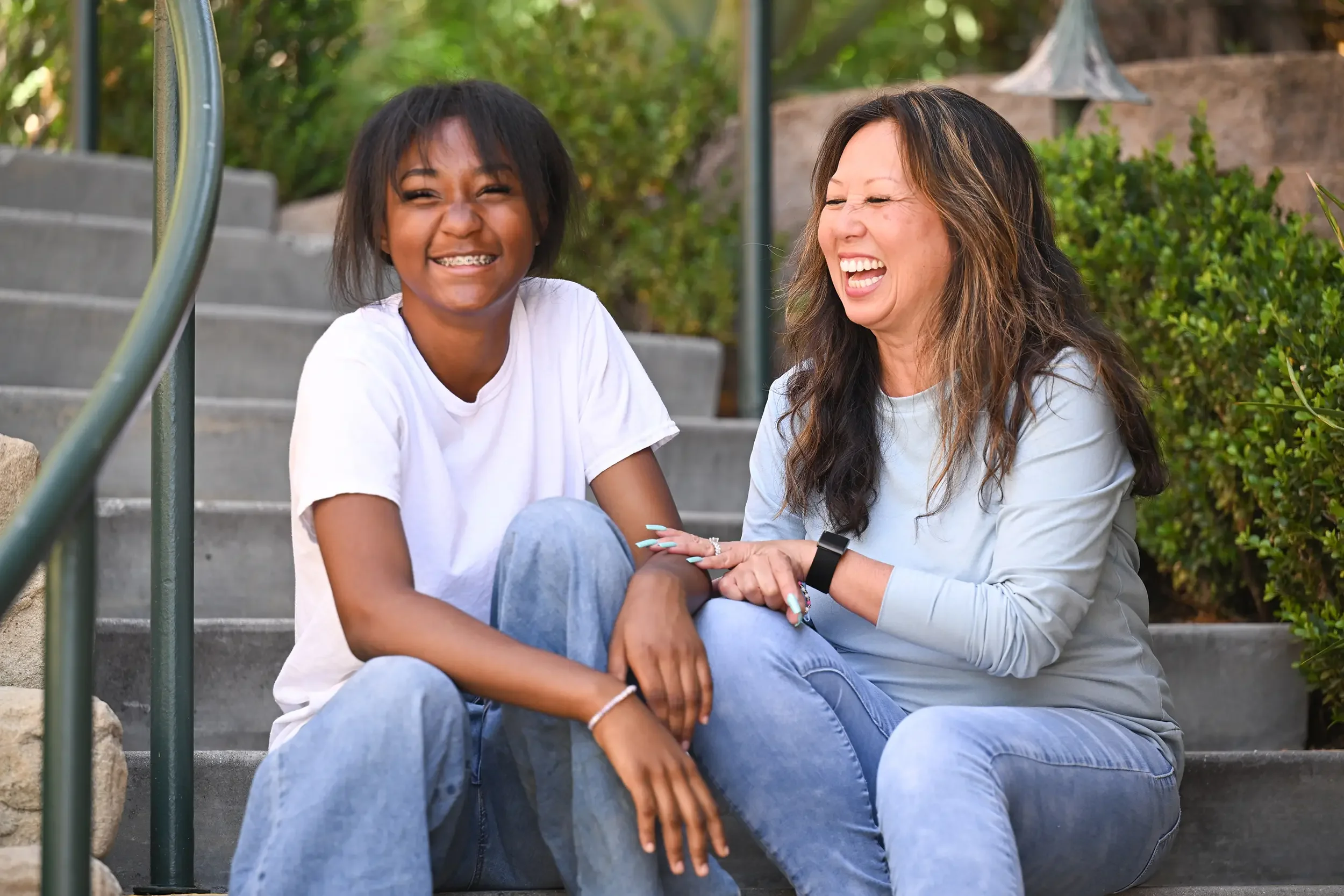 Two women sitting on outdoor stairs, smiling and laughing together, surrounded by greenery.