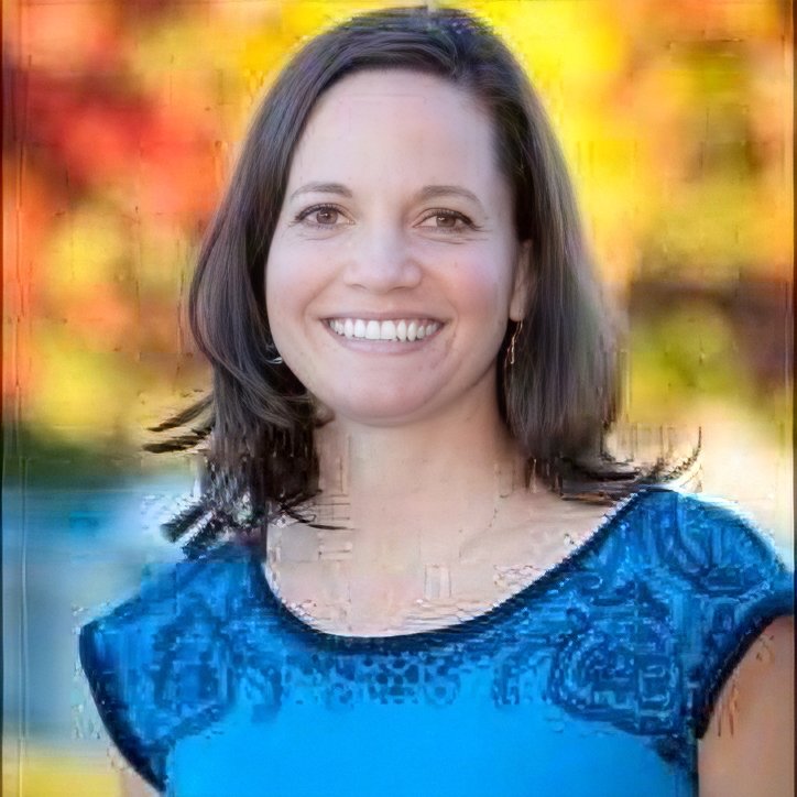 A woman with shoulder-length dark hair smiling outdoors with colorful autumn trees in the background.