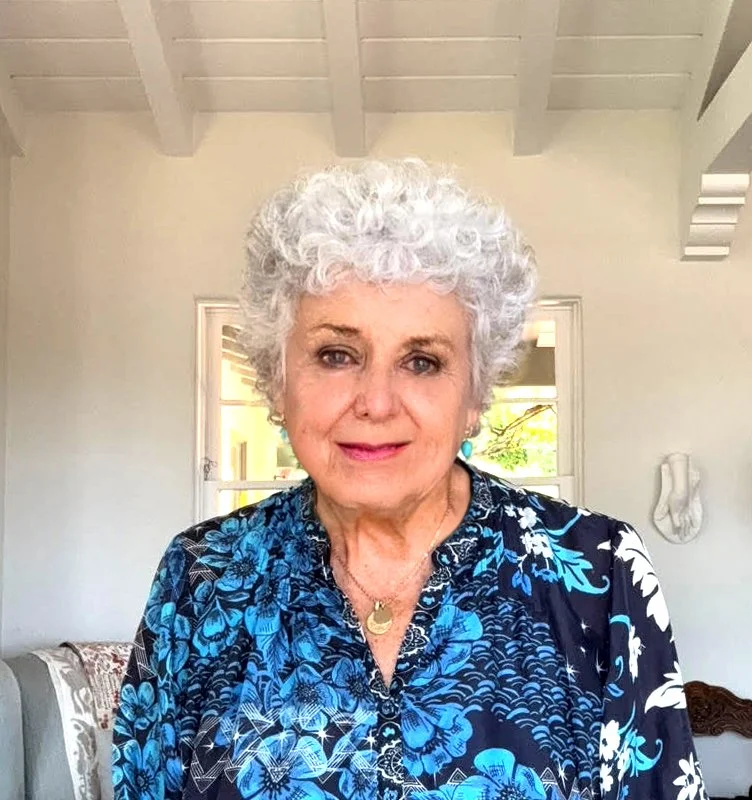 An elderly woman with curly white hair, wearing a blue floral blouse and jewelry, smiling in a bright room with windows and white walls.
