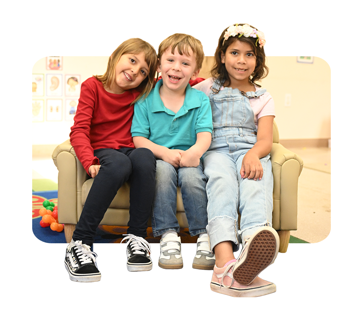 Three children sitting on a beige couch, smiling and posing for the camera. They are in a room with children's artwork and colorful toys in the background.