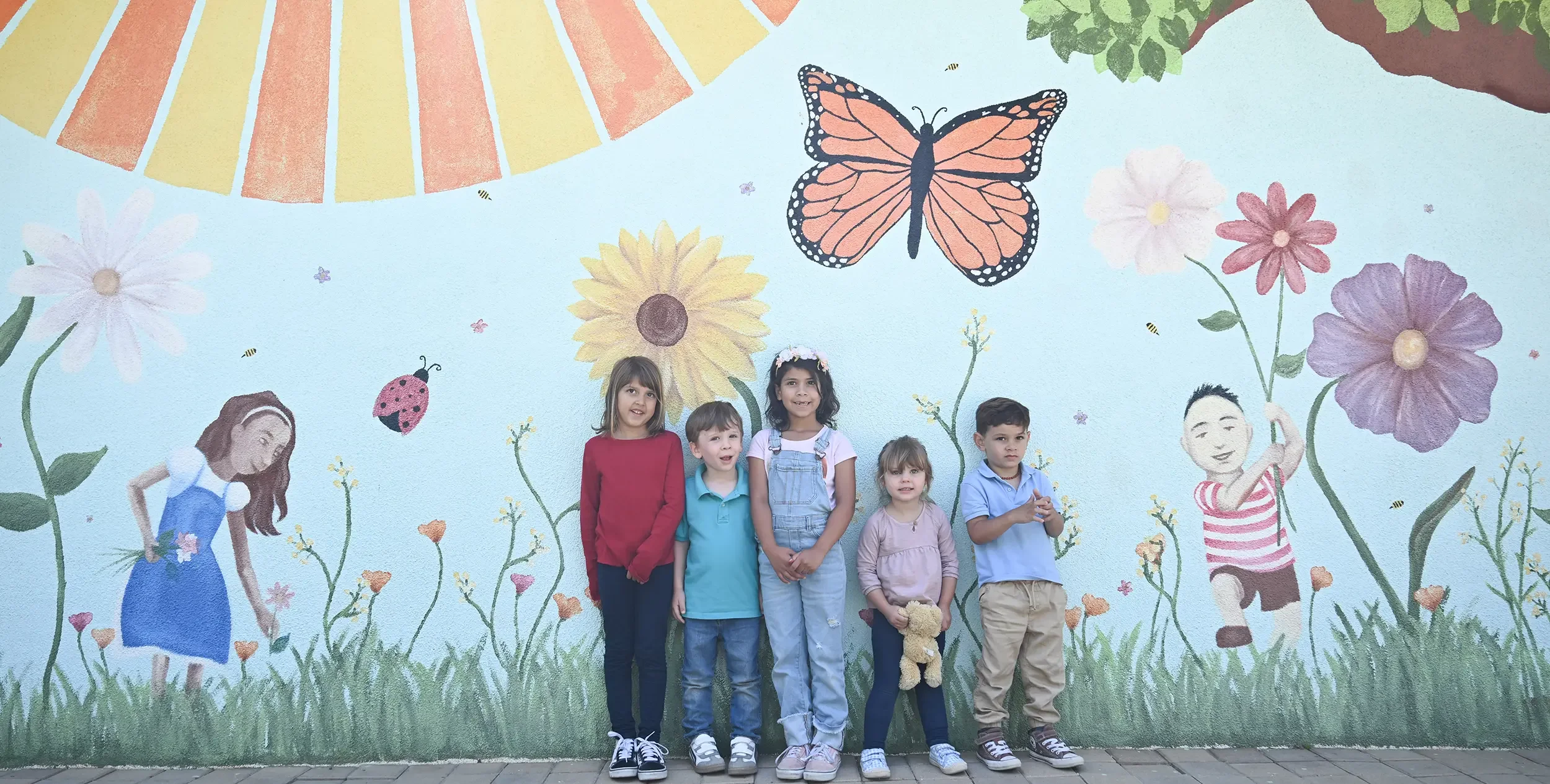 Five children standing in front of a colorful mural featuring large flowers, butterflies, and cartoon characters.