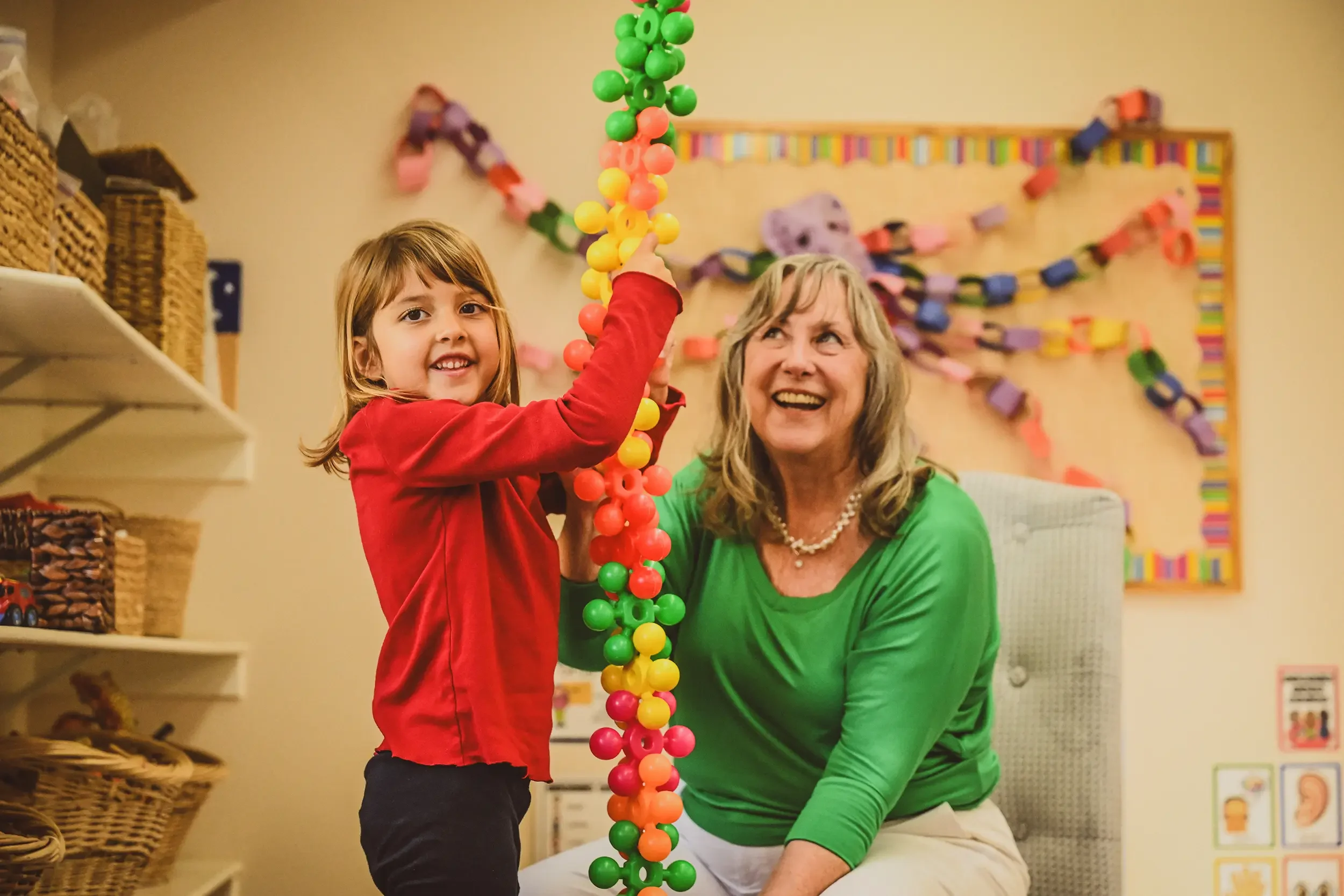 A young girl and an adult woman assembling a large colorful paper chain with pink, yellow, green, and orange balls in a classroom decorated with a multicolored paper chain garland and educational posters.