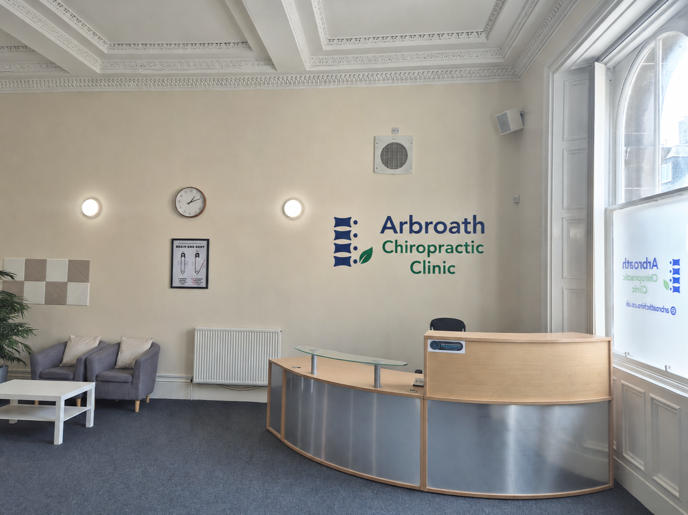 Reception area of Arbroath Chiropractic Clinic with front desk, two gray chairs with pillows, a small white table, and a large window. The clinic's logo and name are on the wall, with a clock, two circular wall lights, and a framed diagram on the wall.