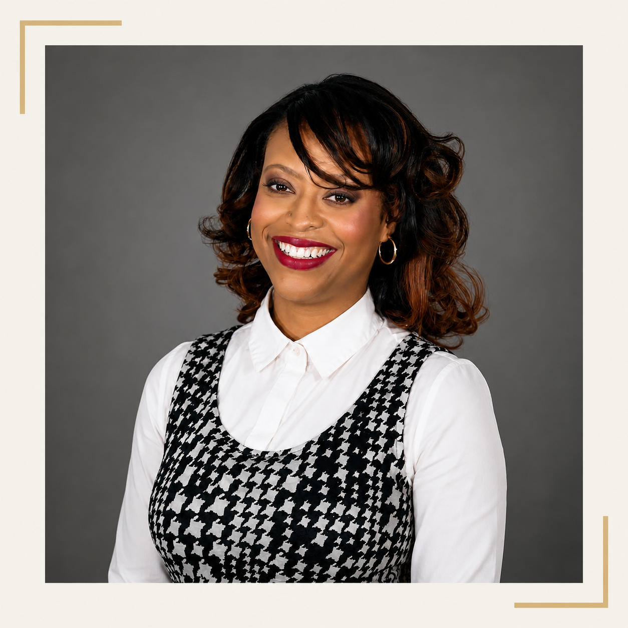 A woman with curly black and brown hair, wearing a white blouse and black and white houndstooth patterned vest, smiling against a gray background.