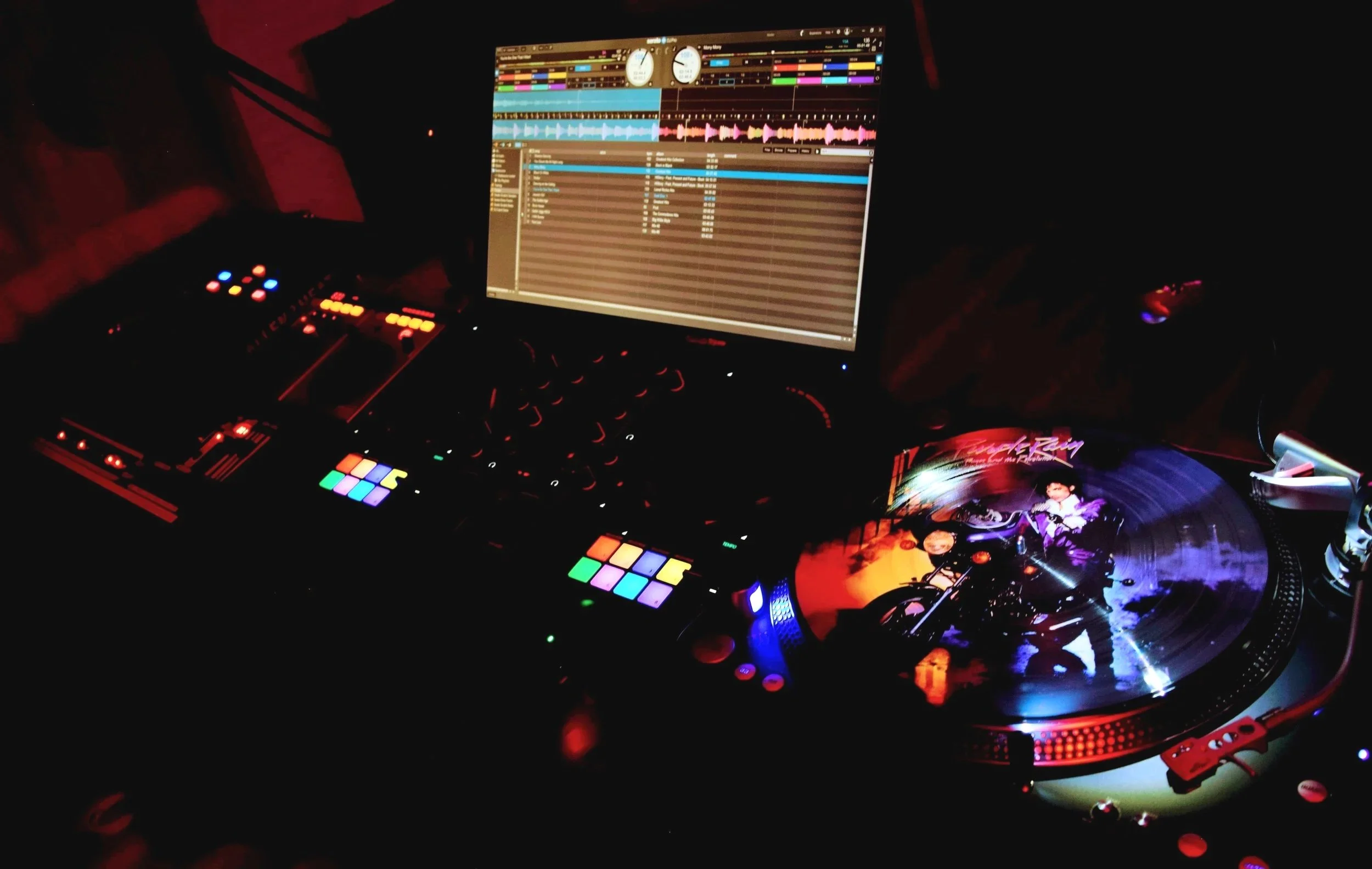 A DJ setup in a dark room with various equipment including a computer monitor showing a music mixing program, a turntable playing vinyl records, and colorful illuminated pads and controls.