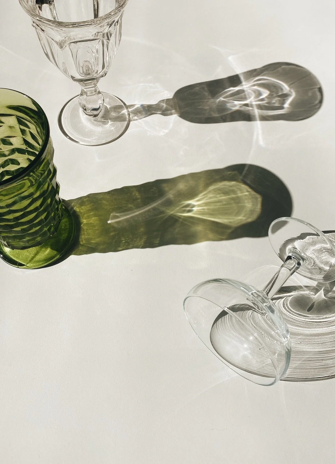 Two empty wine glasses and a green textured vase on a white surface, with sunlight creating reflections and shadows.