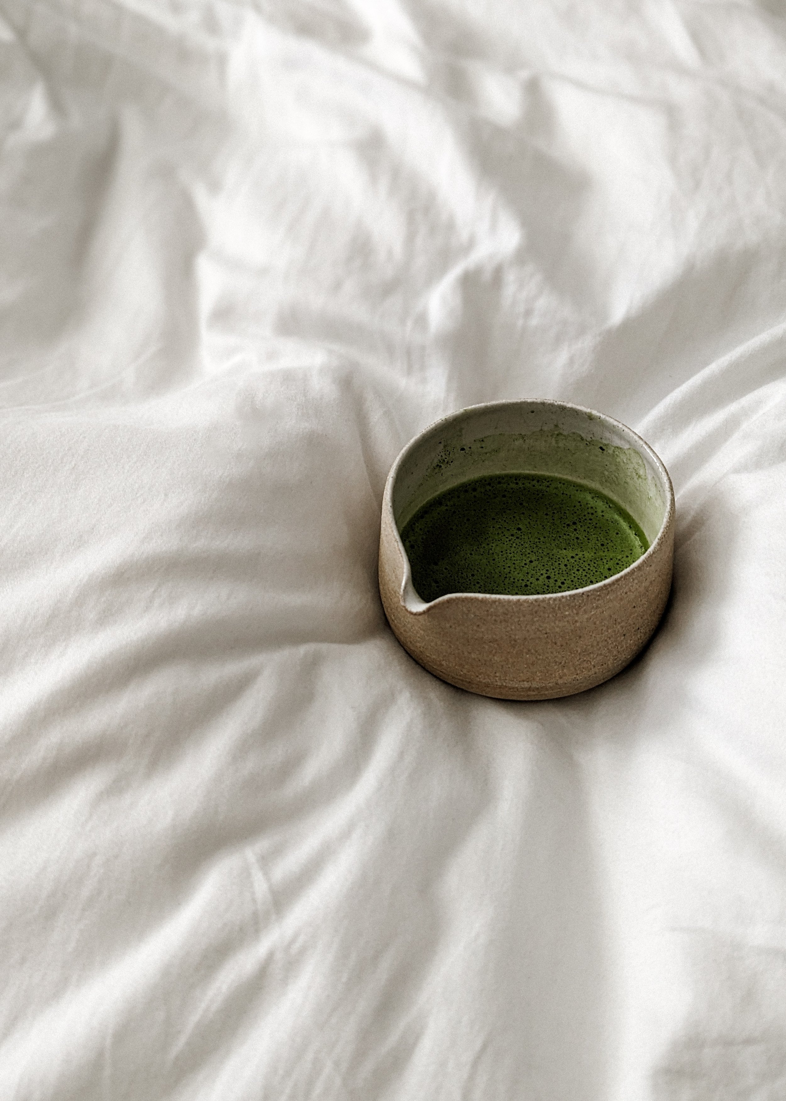 A cup of green matcha tea on a crumpled white bedsheet.