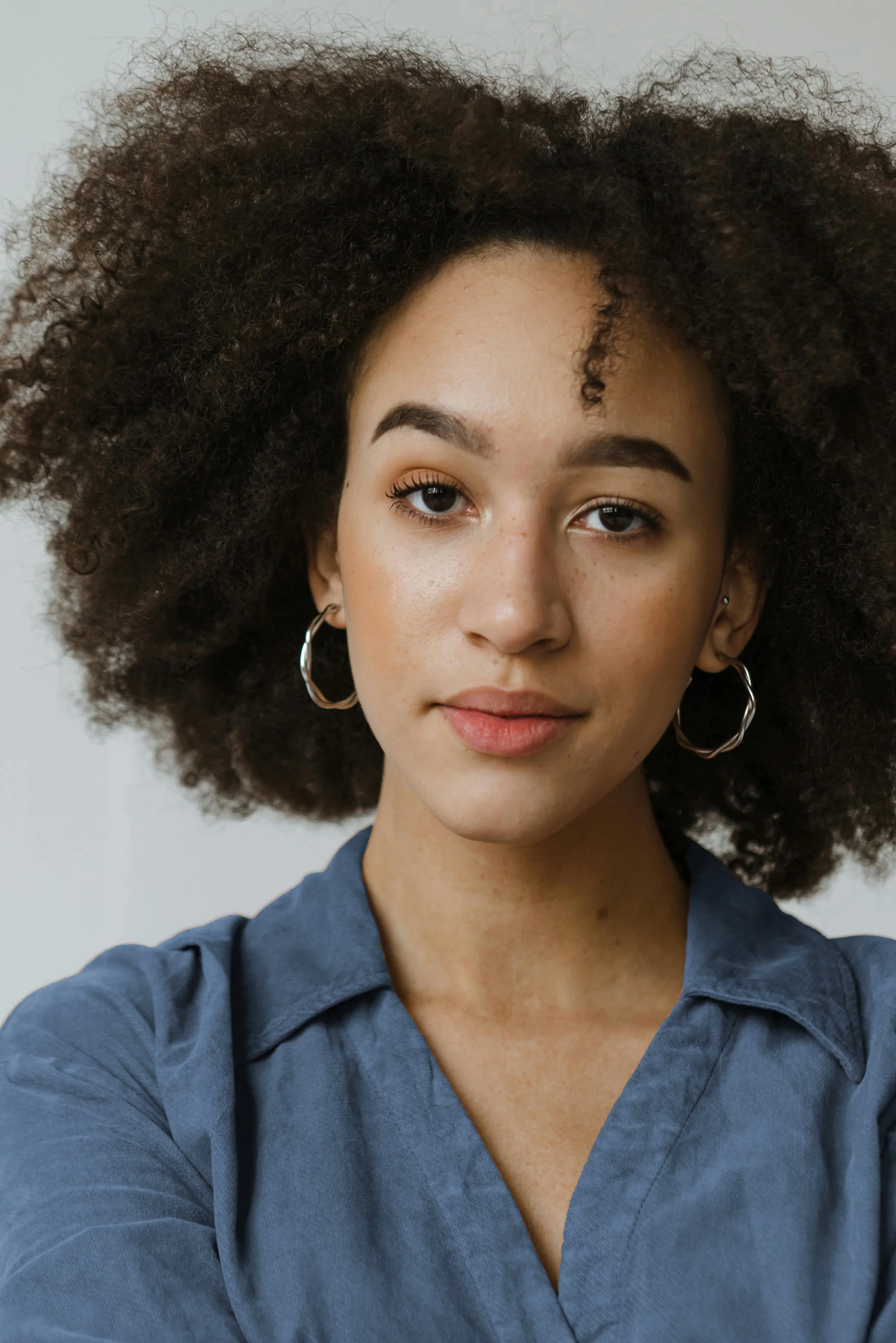 Close-up of a young woman with natural curly hair, wearing hoop earrings and a denim shirt, smiling softly at the camera.