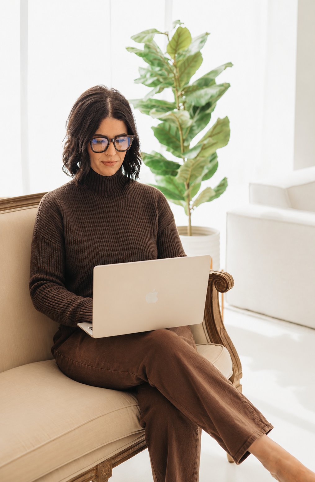 A woman with dark wavy hair and glasses working on a silver MacBook laptop while sitting on a vintage cream-colored sofa in a bright room with white walls and a large green leafy plant in a white pot behind her.