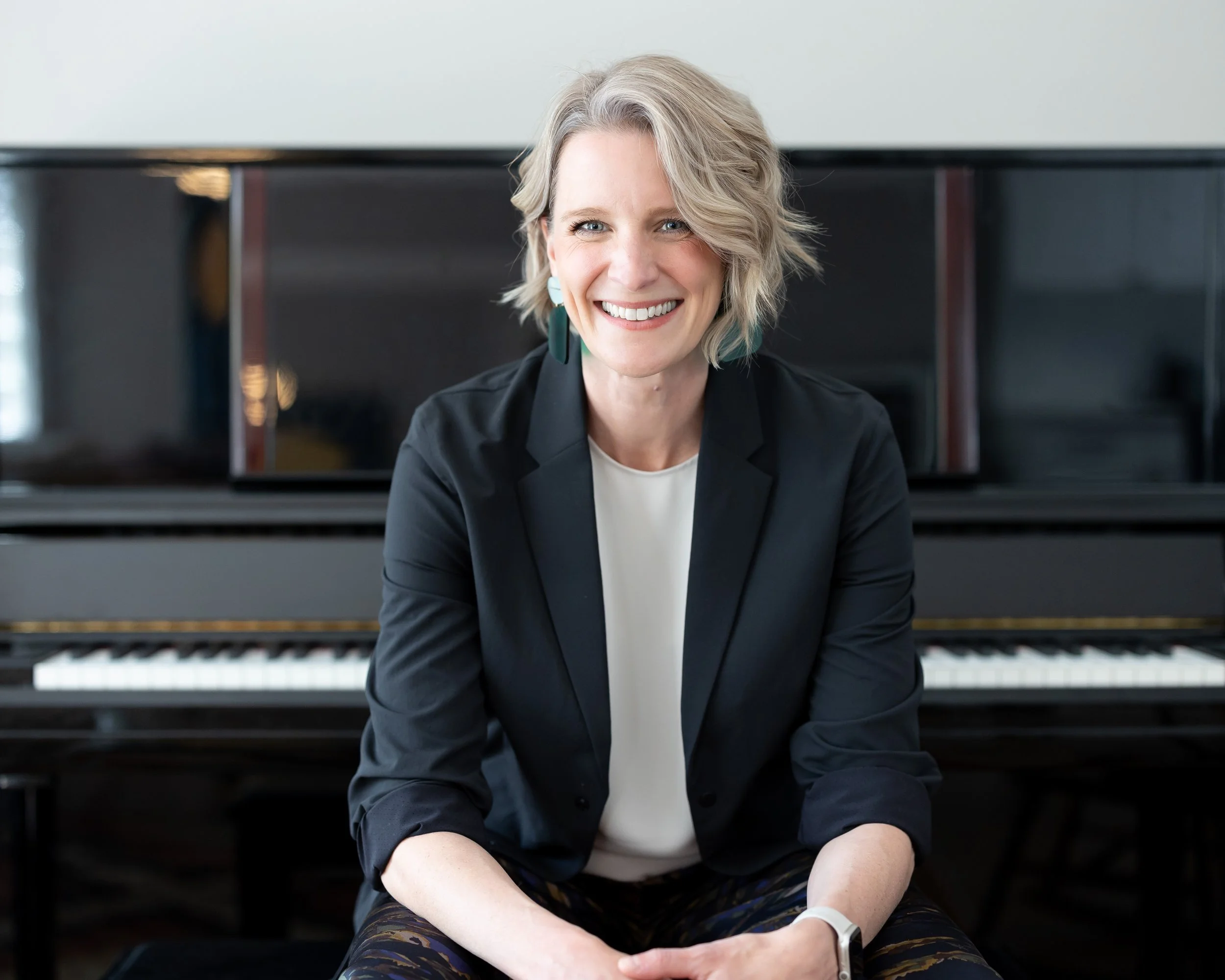 A woman with short blonde hair, wearing a black blazer and white top, sitting in front of a black piano and smiling at the camera.