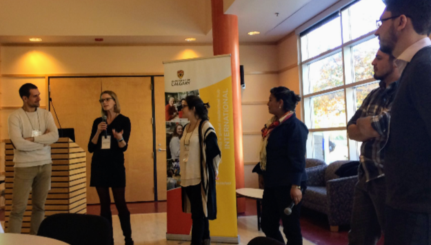 A woman with glasses holding a microphone speaking to a group of people indoors, with a banner in the background and large windows letting in natural light.