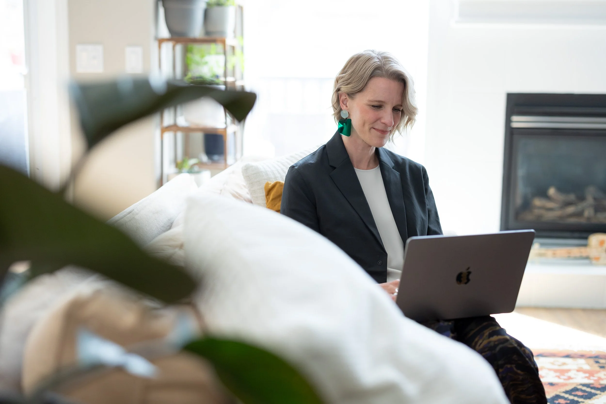 A woman with short blonde hair, wearing a dark blazer and statement earrings, is sitting on a white couch, working on a silver laptop, in a bright room with a patterned rug.
