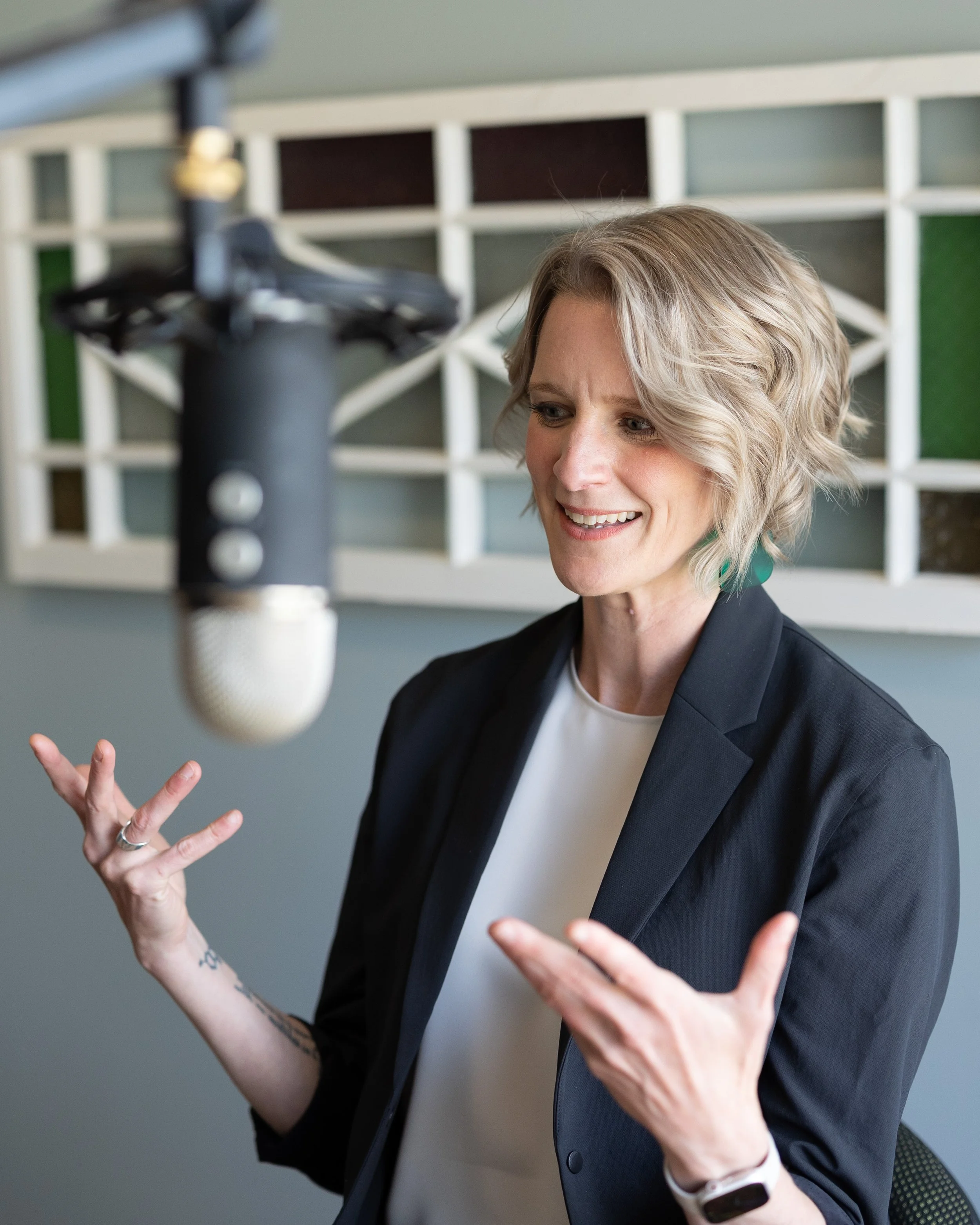 A woman with short blonde curly hair, wearing a black blazer and white shirt, gestures with her hands while speaking into a camera in front of her. The background features a wall with a geometric pattern of multicolored squares.