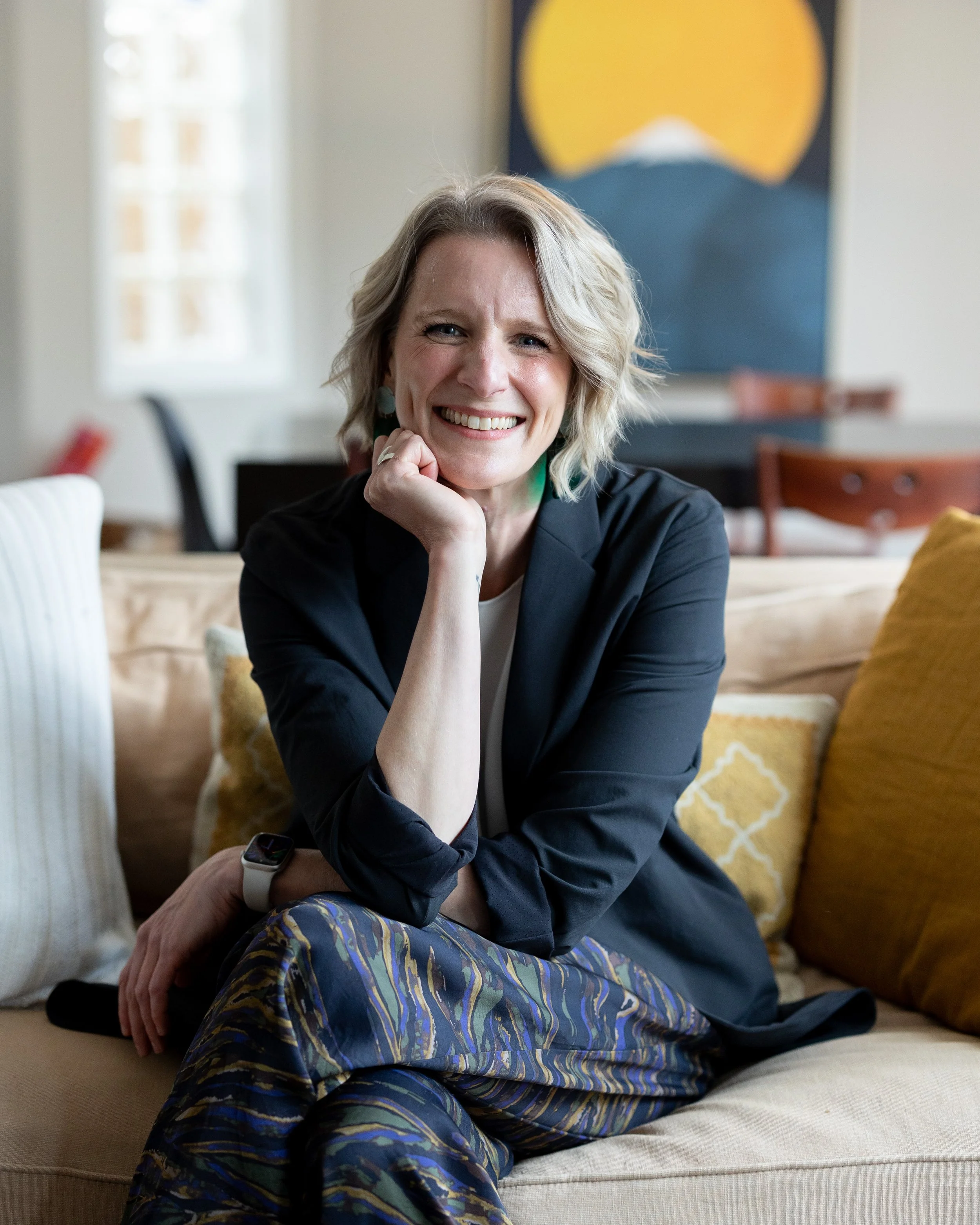 A smiling woman with blonde, curly hair sitting inside a cozy living room on a beige couch. She is wearing a black blazer and colorful patterned pants. There are decorative pillows behind her and a piece of modern art on the wall in the background.