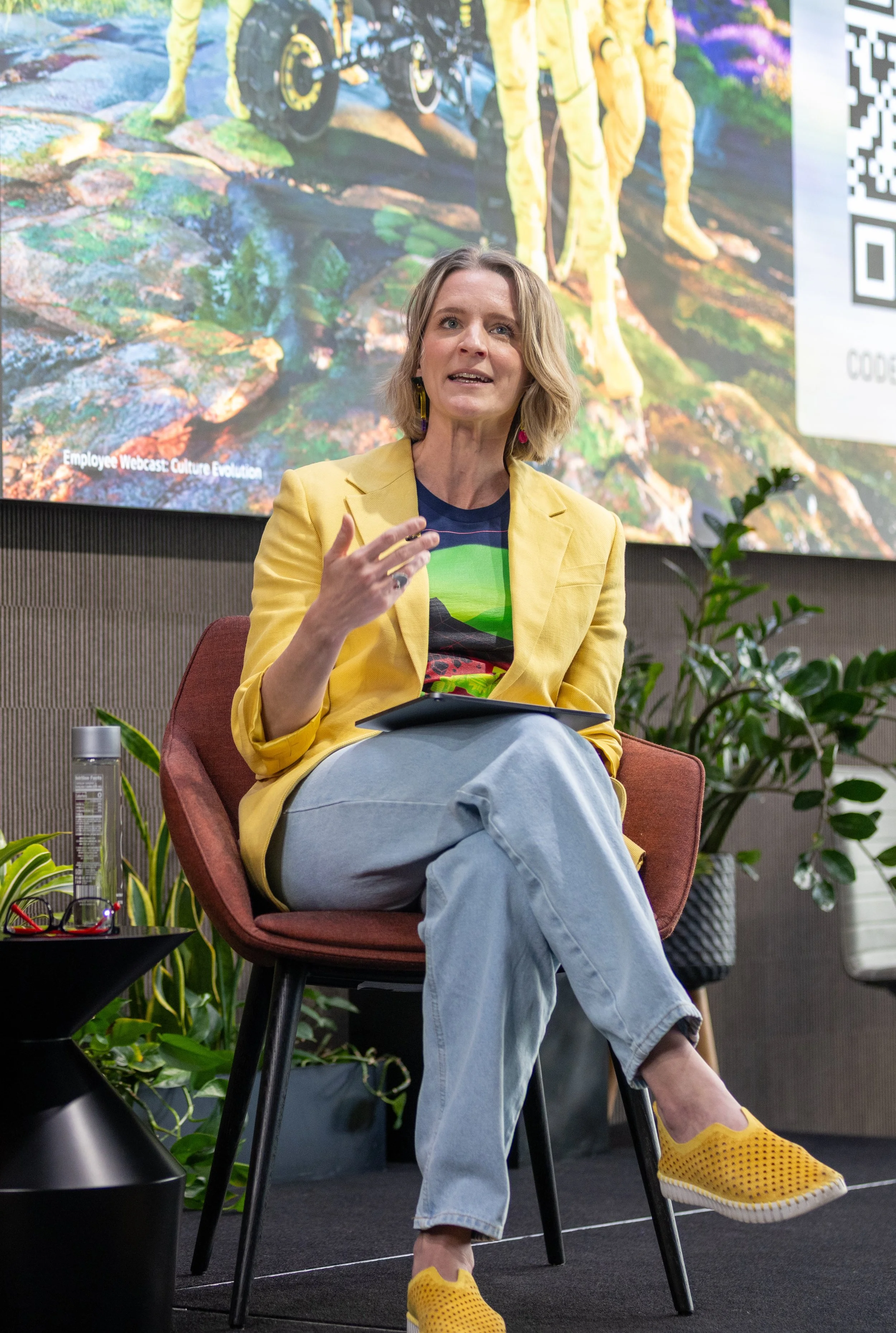 A woman seated in a chair, speaking with her hand raised. She has shoulder-length blond hair and is wearing a yellow blazer, a printed T-shirt, light blue jeans, and yellow sneakers. Behind her, there is a large colorful mural of a landscape and a digital screen.