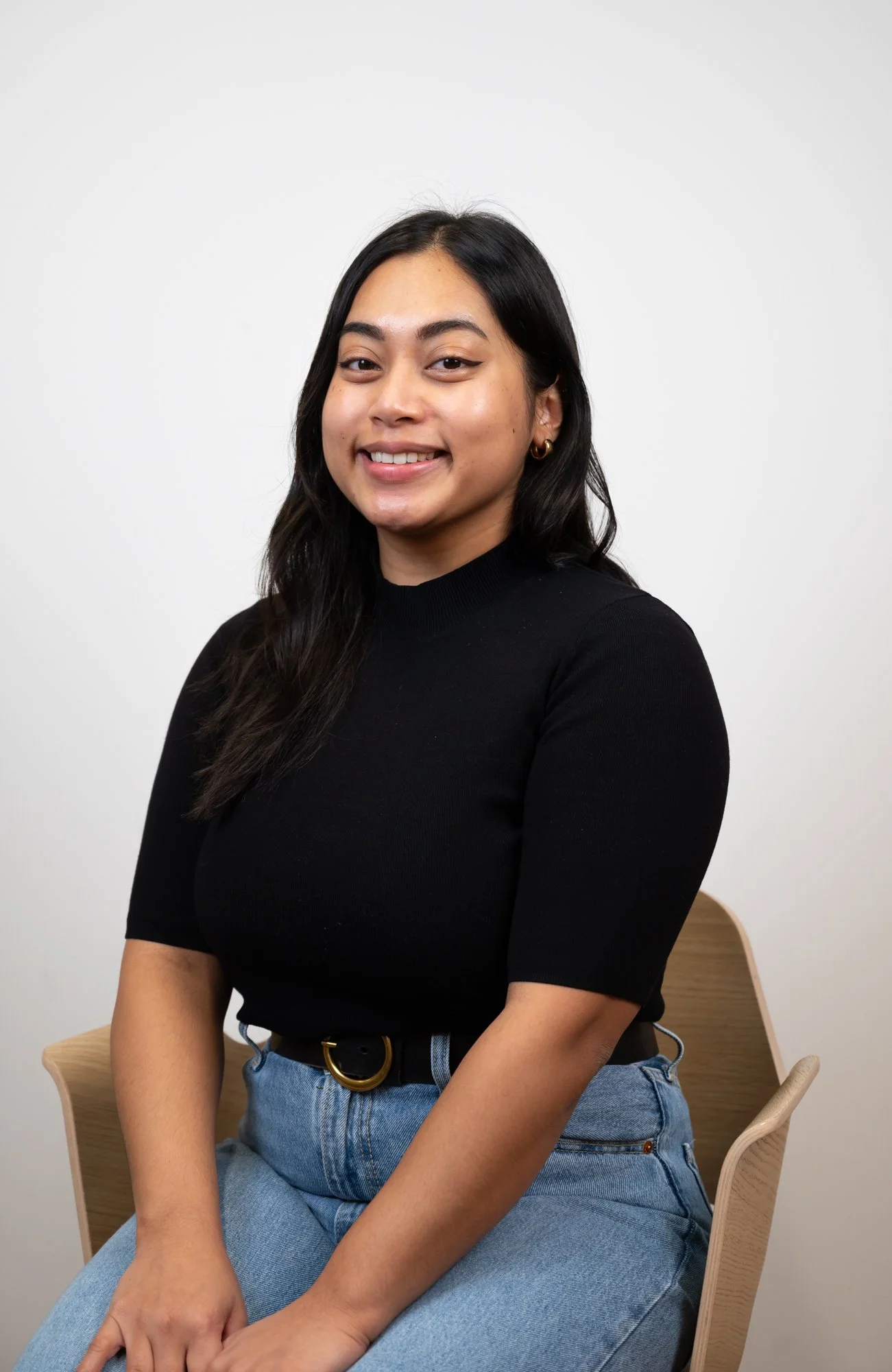 A woman with long black hair and hoop earrings, smiling while sitting on a wooden chair against a plain white wall, wearing a black top and light blue jeans.