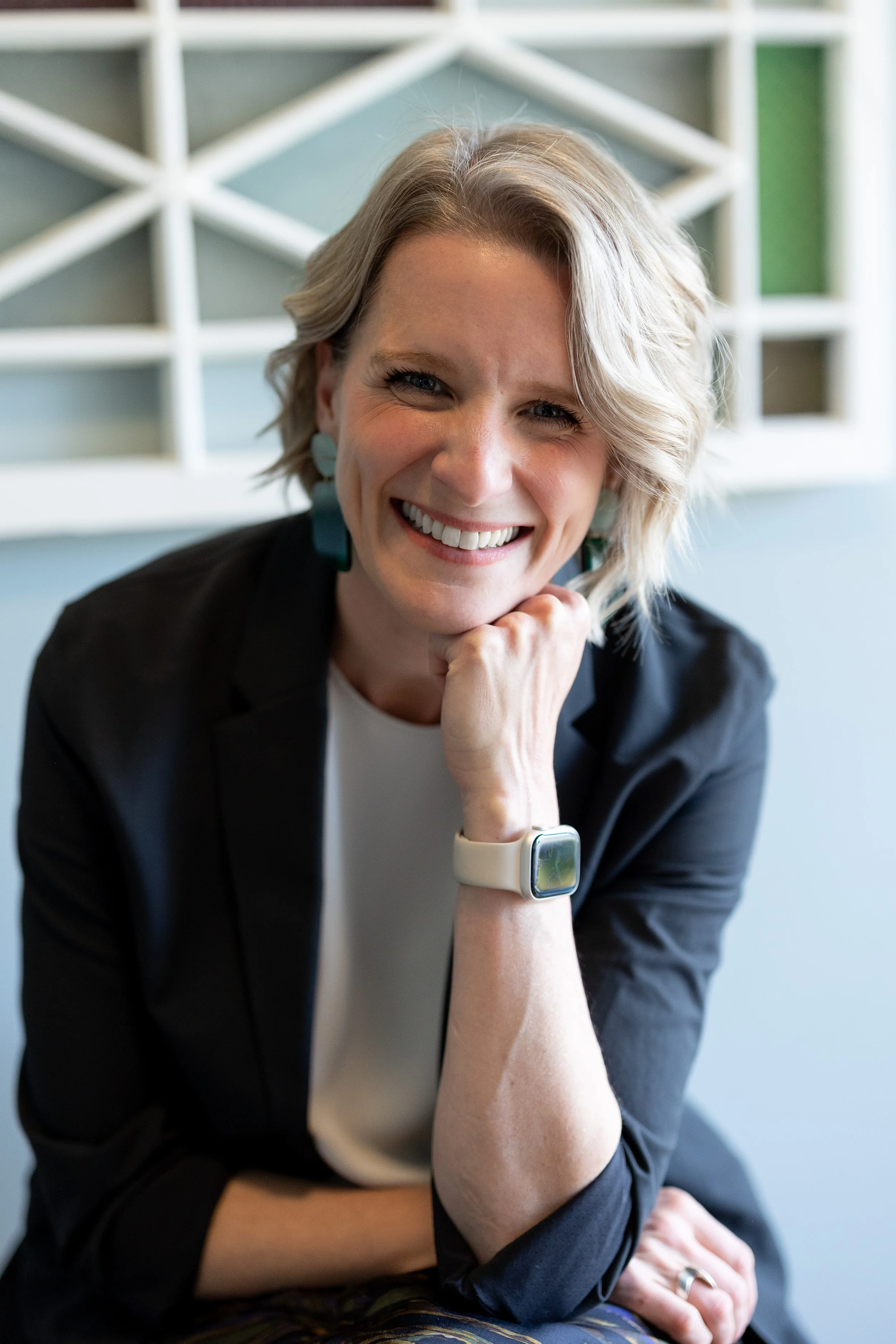 A woman smiling while resting her chin on her hand, wearing a black blazer, white shirt, and a smartwatch on her wrist, with a geometric wall pattern in the background.