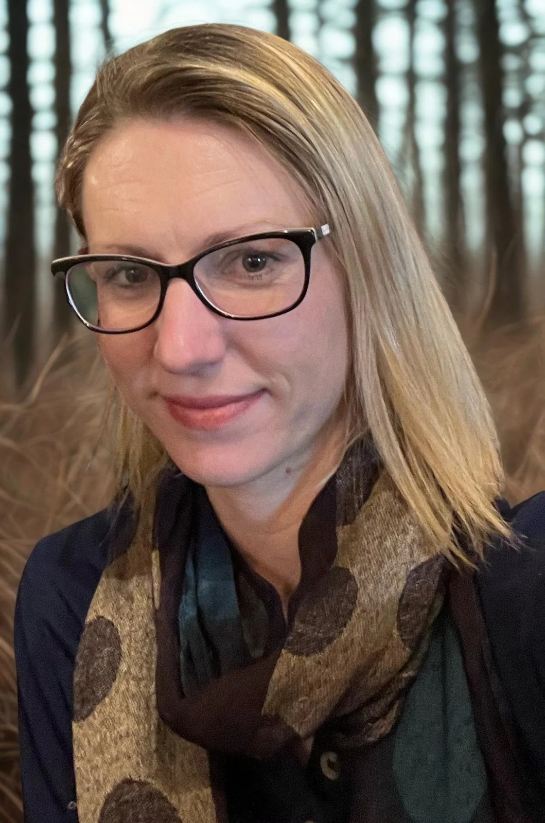 A woman with blonde hair and black glasses smiling outdoors in a wooded area.