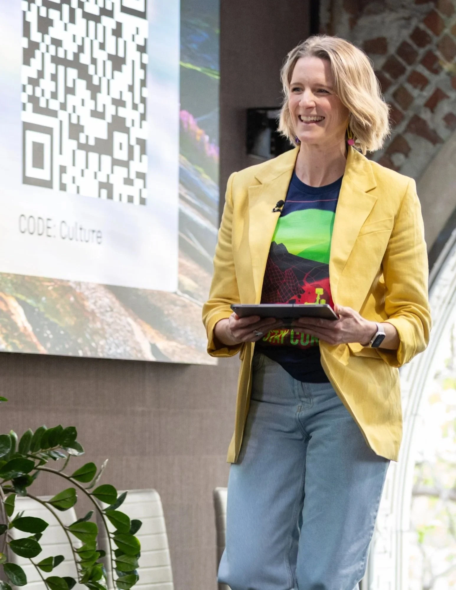 A woman with blonde hair smiling and holding a tablet, standing near a large QR code poster with the text "CODE: CULTURE" in an indoor setting with a plant and chairs.