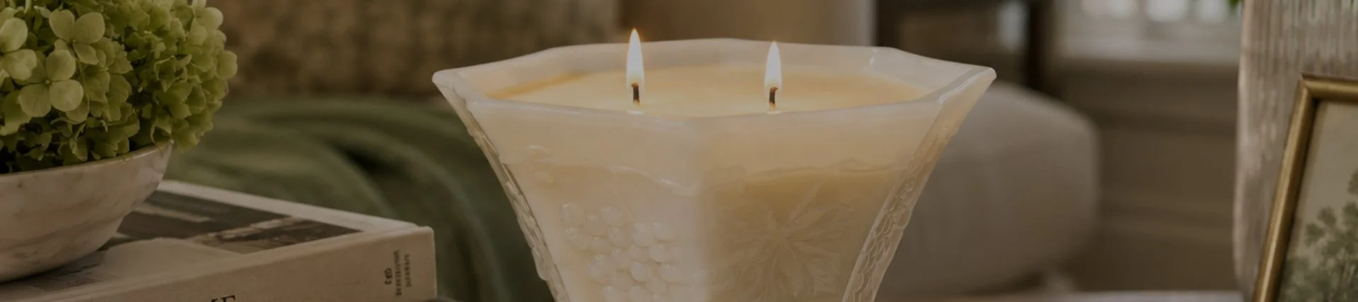 A beige candle with two burning wicks in a clear, decorative glass holder on a table, with a book and a plant nearby.