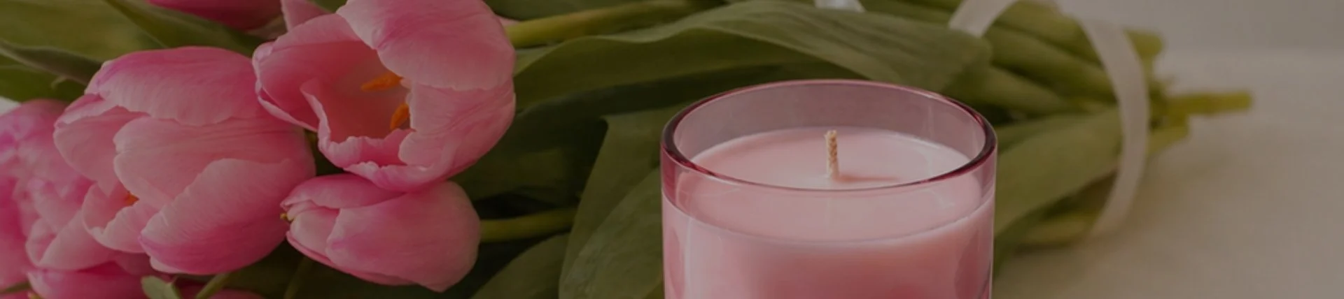 Pink candle with a wick in front of a bouquet of pink tulips with green leaves.