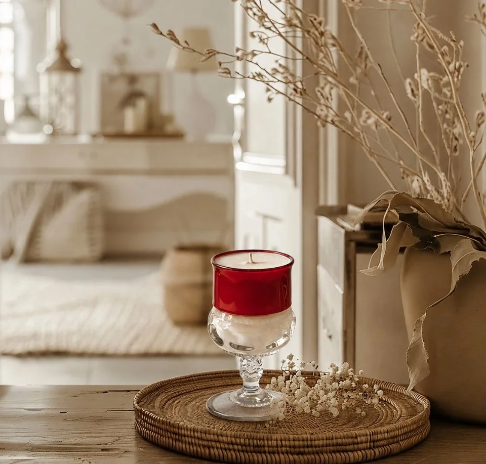 A red and clear glass candle holder on woven tray with dried flowers, beside a large vase with dried branches, in a cozy, neutral-toned room.