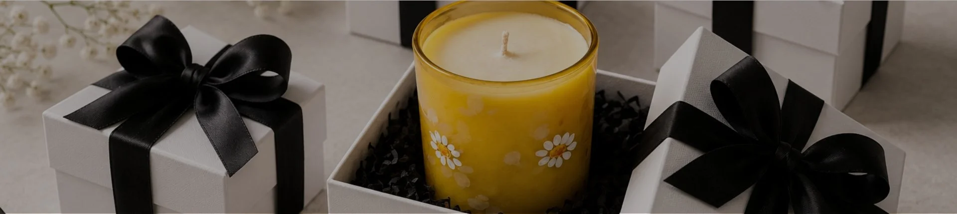 A yellow candle with white daisy floral pattern in a glass container, surrounded by white gift boxes with black ribbon bows, placed on a light-colored surface.