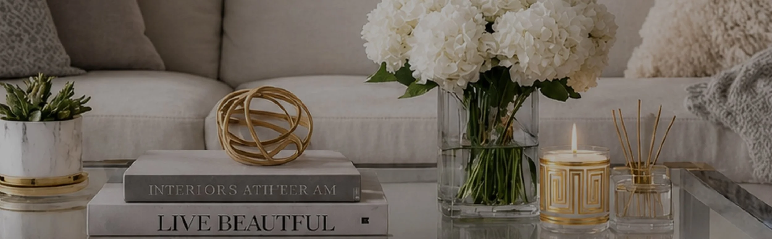Decorative home interior with a marble coffee table, white hydrangea flowers in a glass vase, a gold-trimmed candle, reed diffuser, a potted succulent plant, and books titled 'Interiors Attheer Am' and 'Live Beautiful' in a cozy living room.