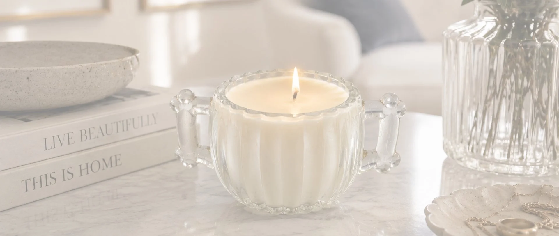 A lit candle in a clear, decorative glass holder on a white marble surface. To the left, two books stacked with titles 'LIVE BEAUTIFULLY' and 'THIS IS HOME.' To the right, a clear, ribbed glass vase with greenery. In the foreground, a small dish with jewelry.