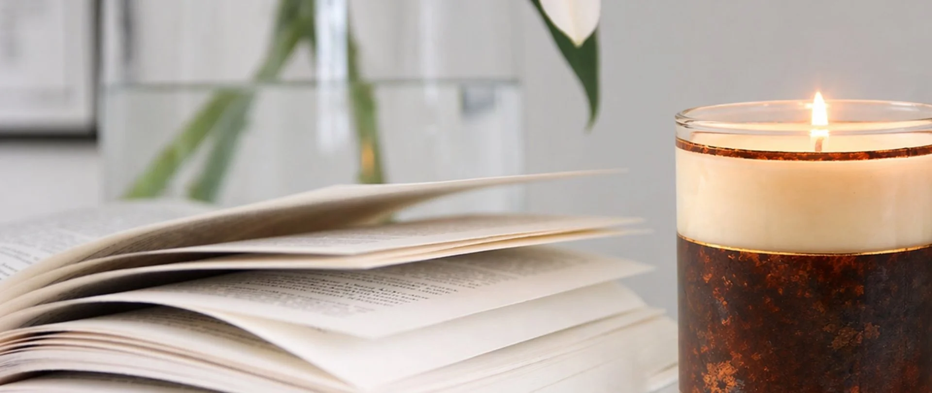 An open book with a few pages turned, a lit candle in a decorative holder, and a vase with flowers in the background.