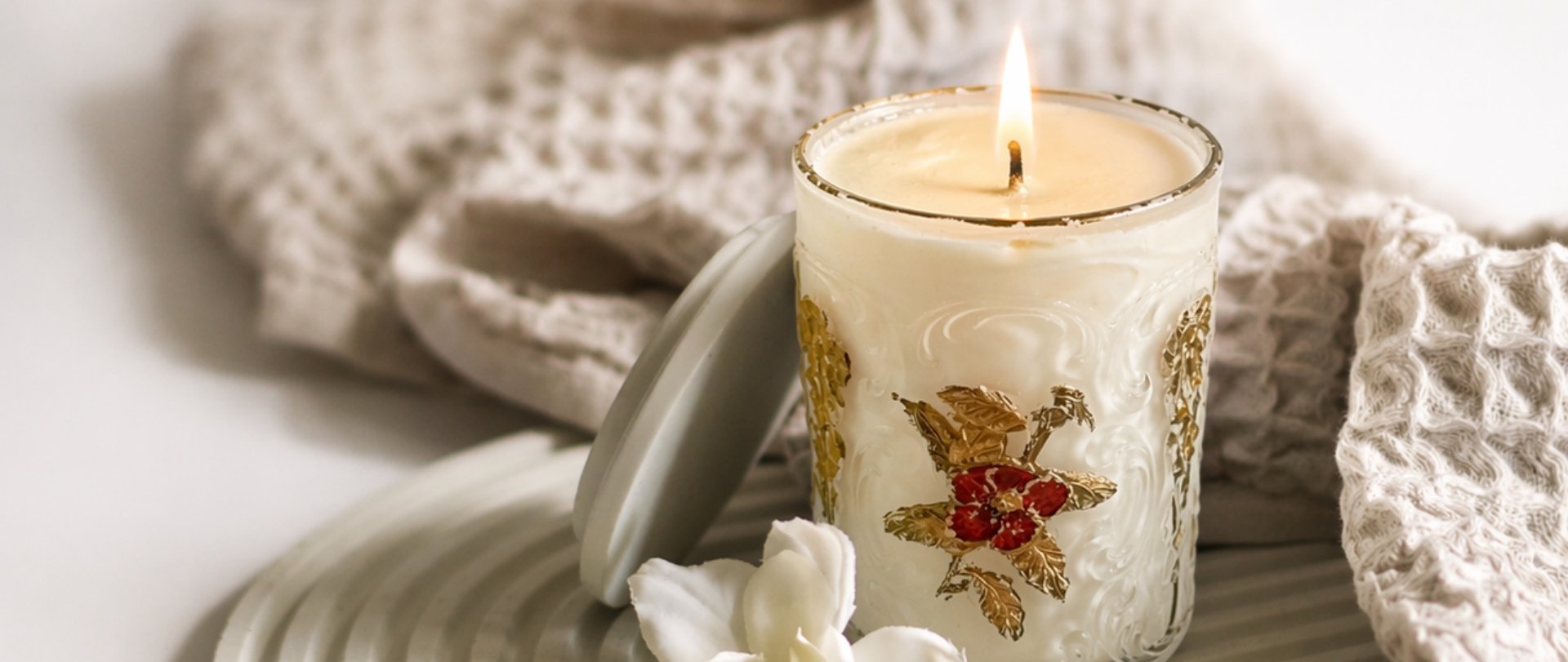 Lit Christmas candle in decorative glass holder with gold and red poinsettia design, surrounded by white knitted fabrics and marshmallows.