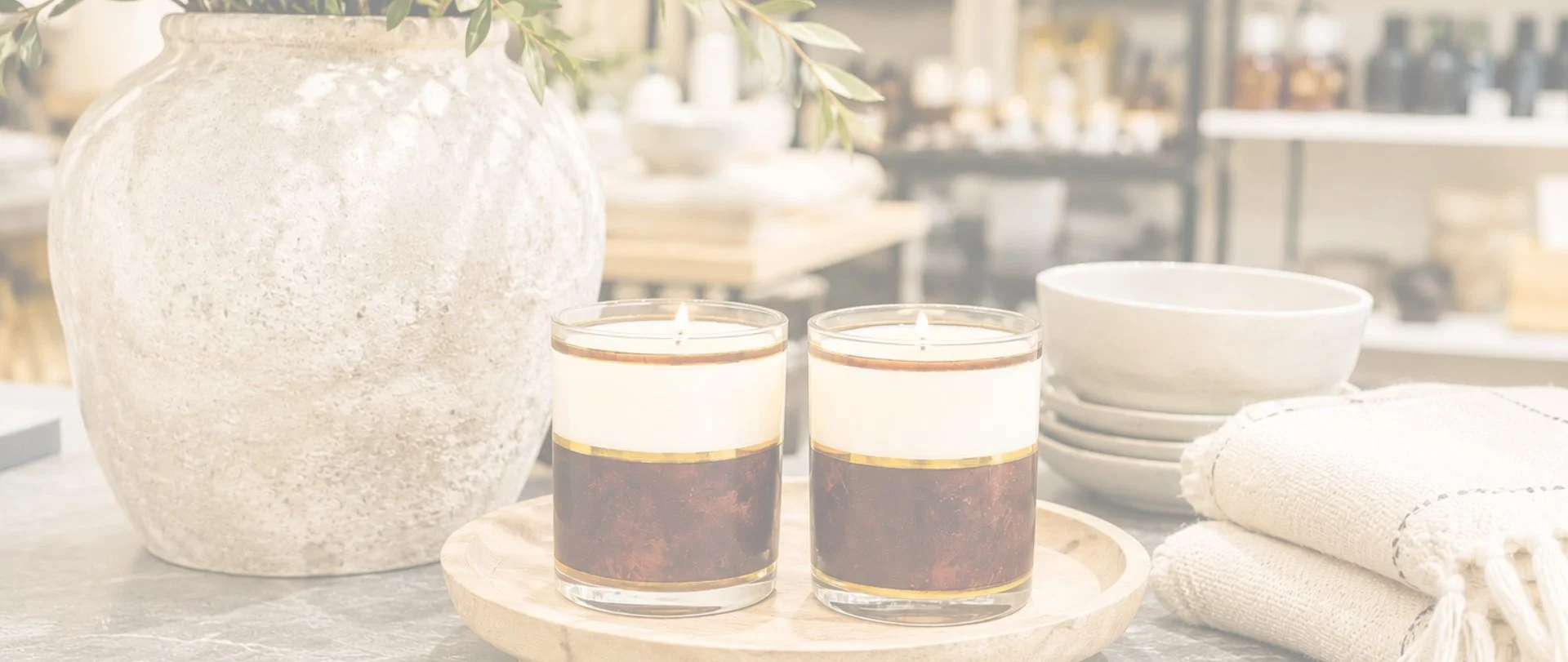 Two lit candles in glass holders on a round wooden tray, surrounded by stacked white bowls, a folded white towel, and a large textured white vase with green leaves, in a bright and airy space.